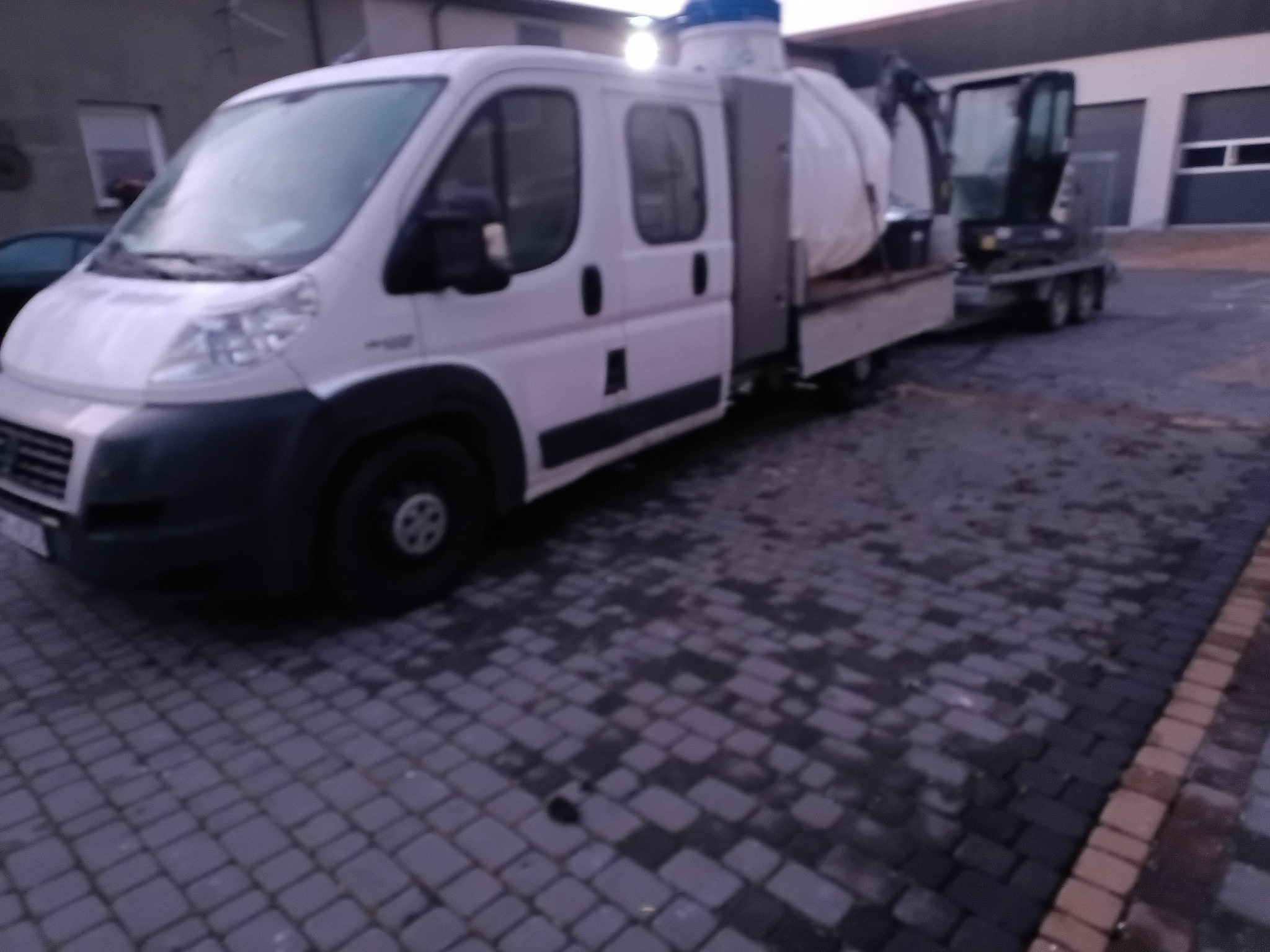 Specjalistyczny pojazd z cysterną i mini koparką na przyczepie, prawdopodobnie do oczyszczania kanalizacji, zaparkowany na brukowanym placu.