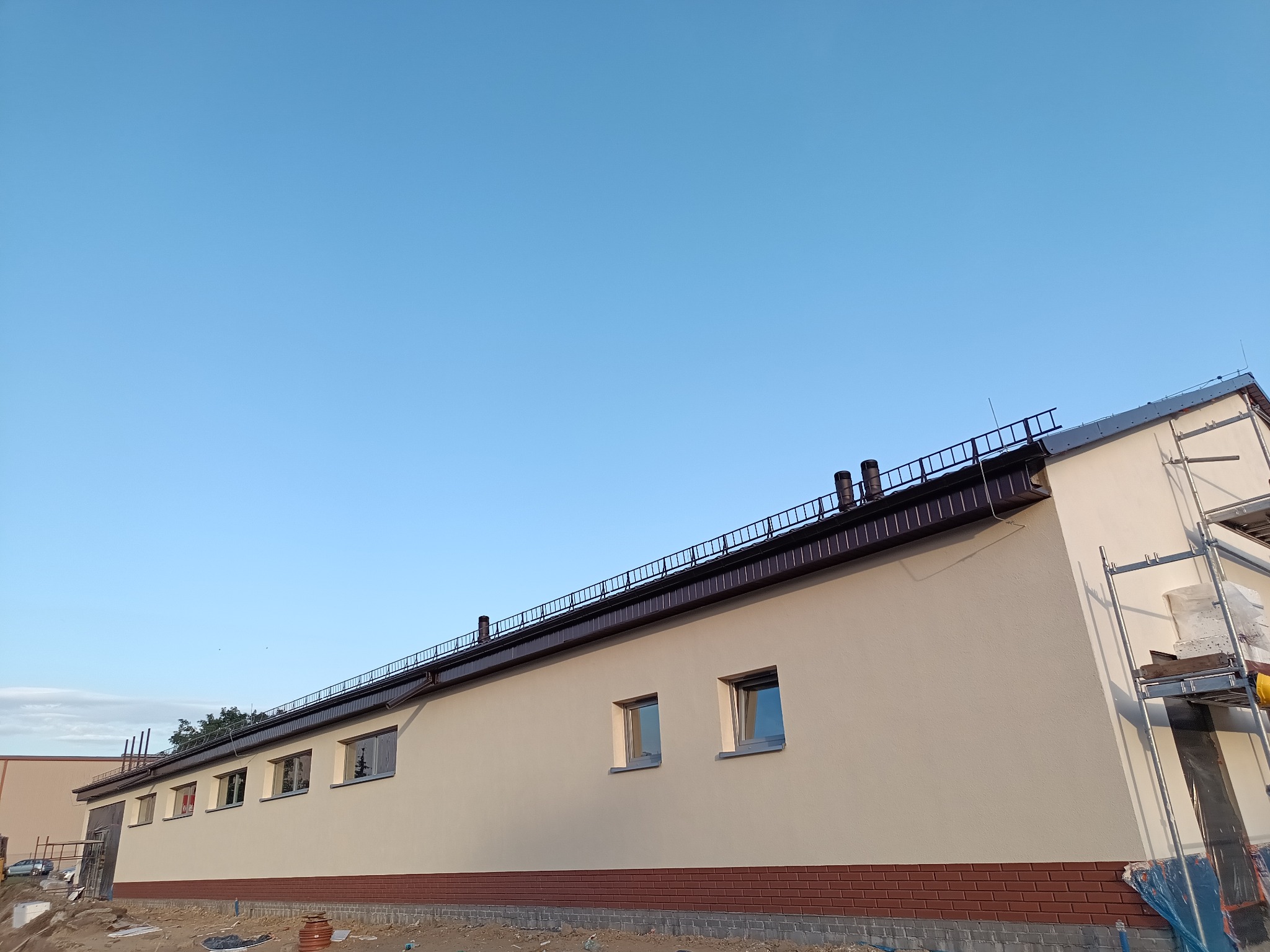 Ocieplona elewacja budynku z ceglaną podmurówką i rzędami okien, widoczna z dołu pod kątem, z rusztowaniem z boku. Jasne niebo w tle.