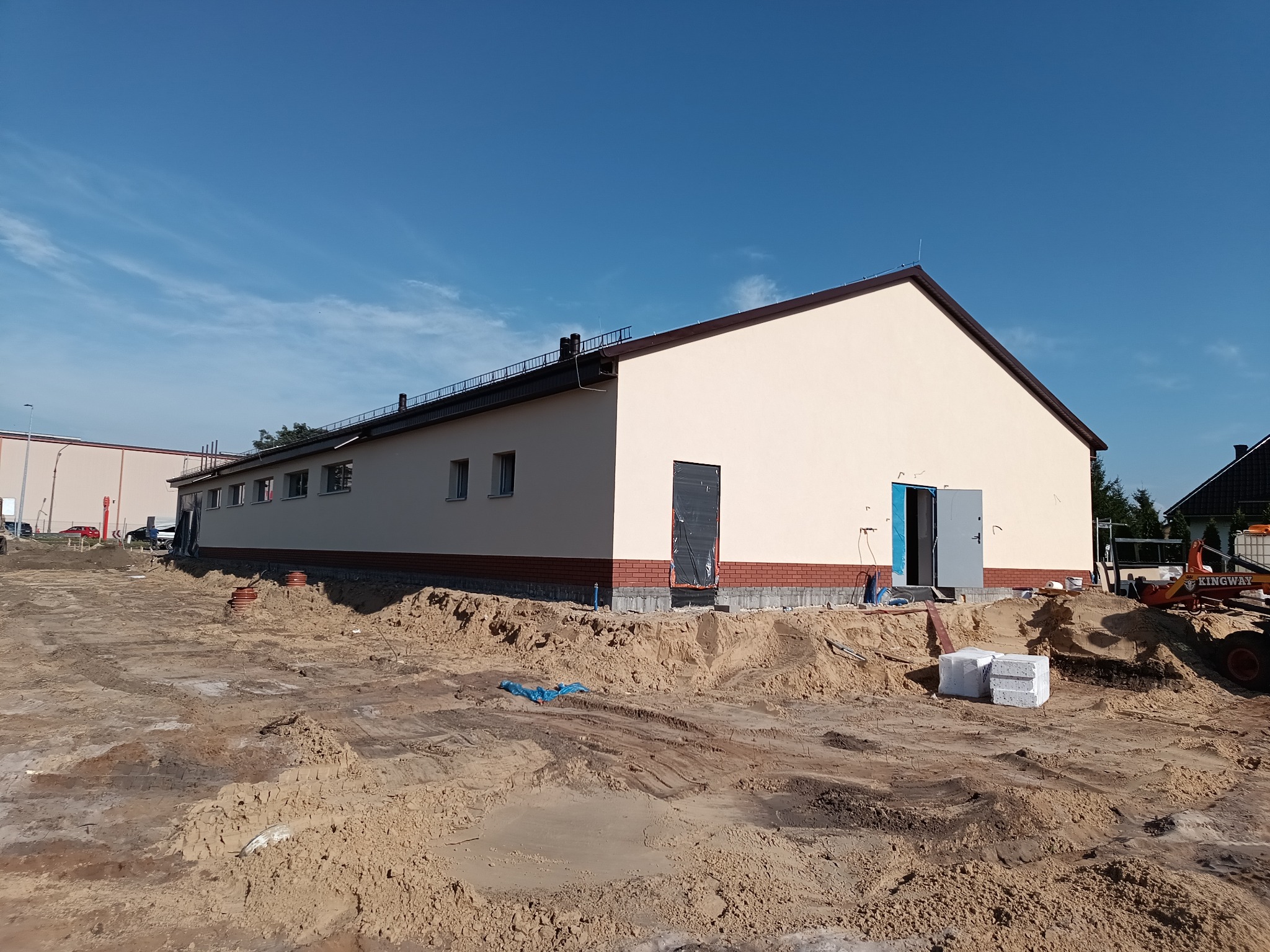 Budynek po ociepleniu elewacji z widocznymi otwartymi drzwiami i przygotowanym terenem wokół. Widoczne materiały budowlane i sprzęt. Jasne niebo.