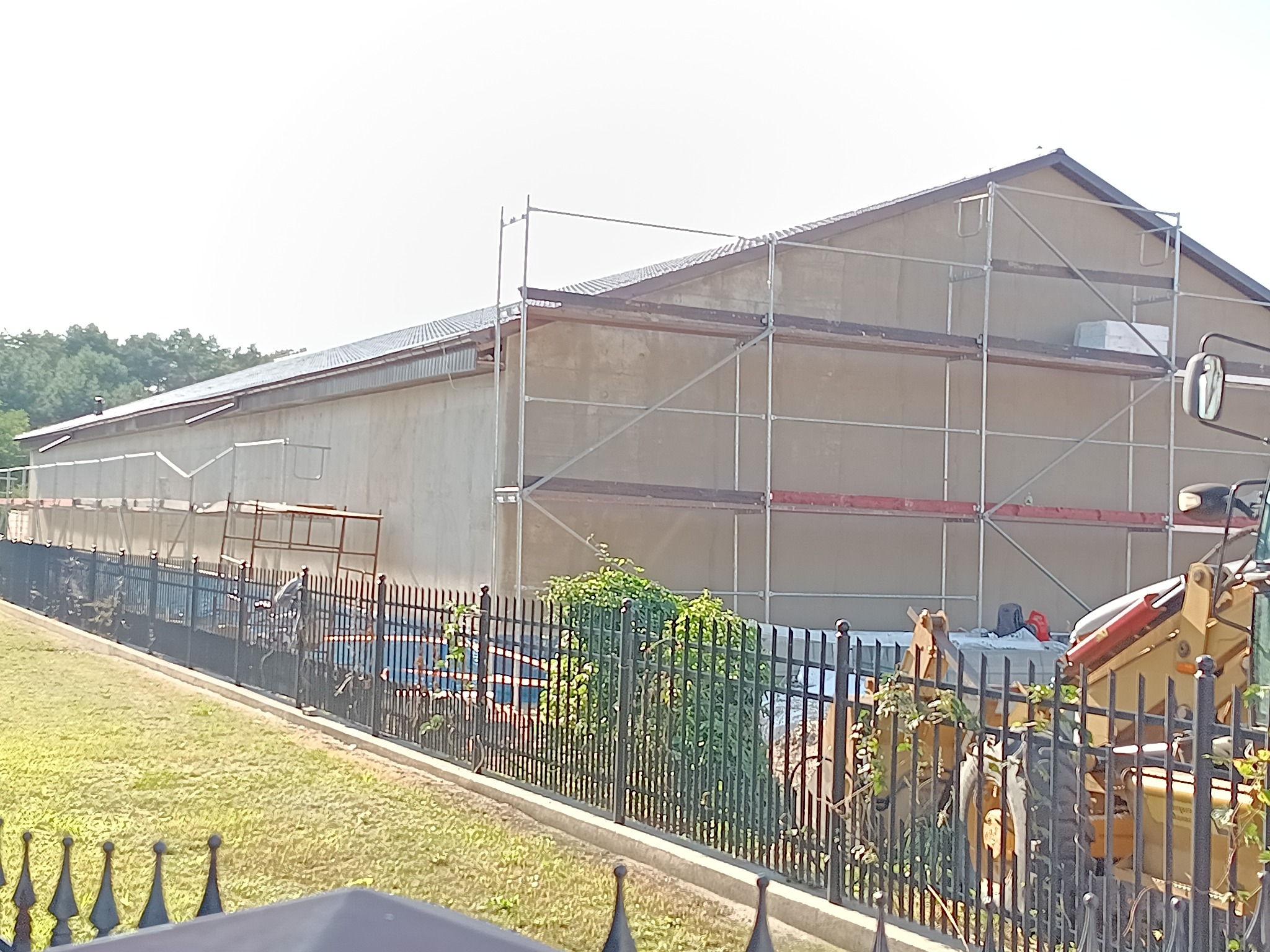 Budynek w trakcie ocieplania, widoczne rusztowania i sprzęt budowlany za ozdobnym ogrodzeniem. Częściowo otynkowana elewacja. Prace w toku, słoneczny dzień.