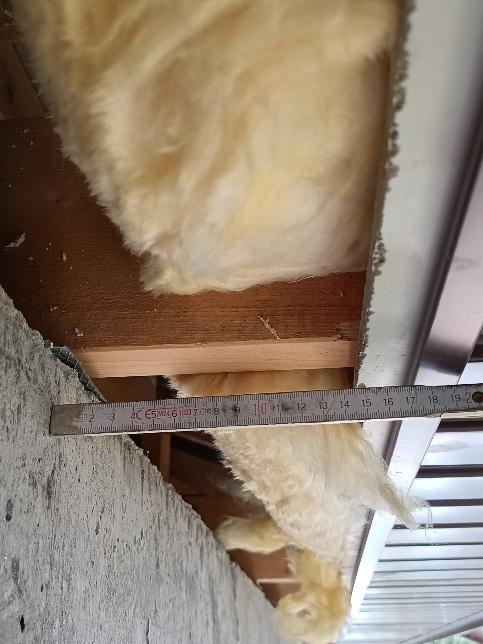 Pomiar grubości izolacji z wełny mineralnej w konstrukcji dachu za pomocą rozłożonej miarki. Widoczne elementy drewniane i metalowe konstrukcji.