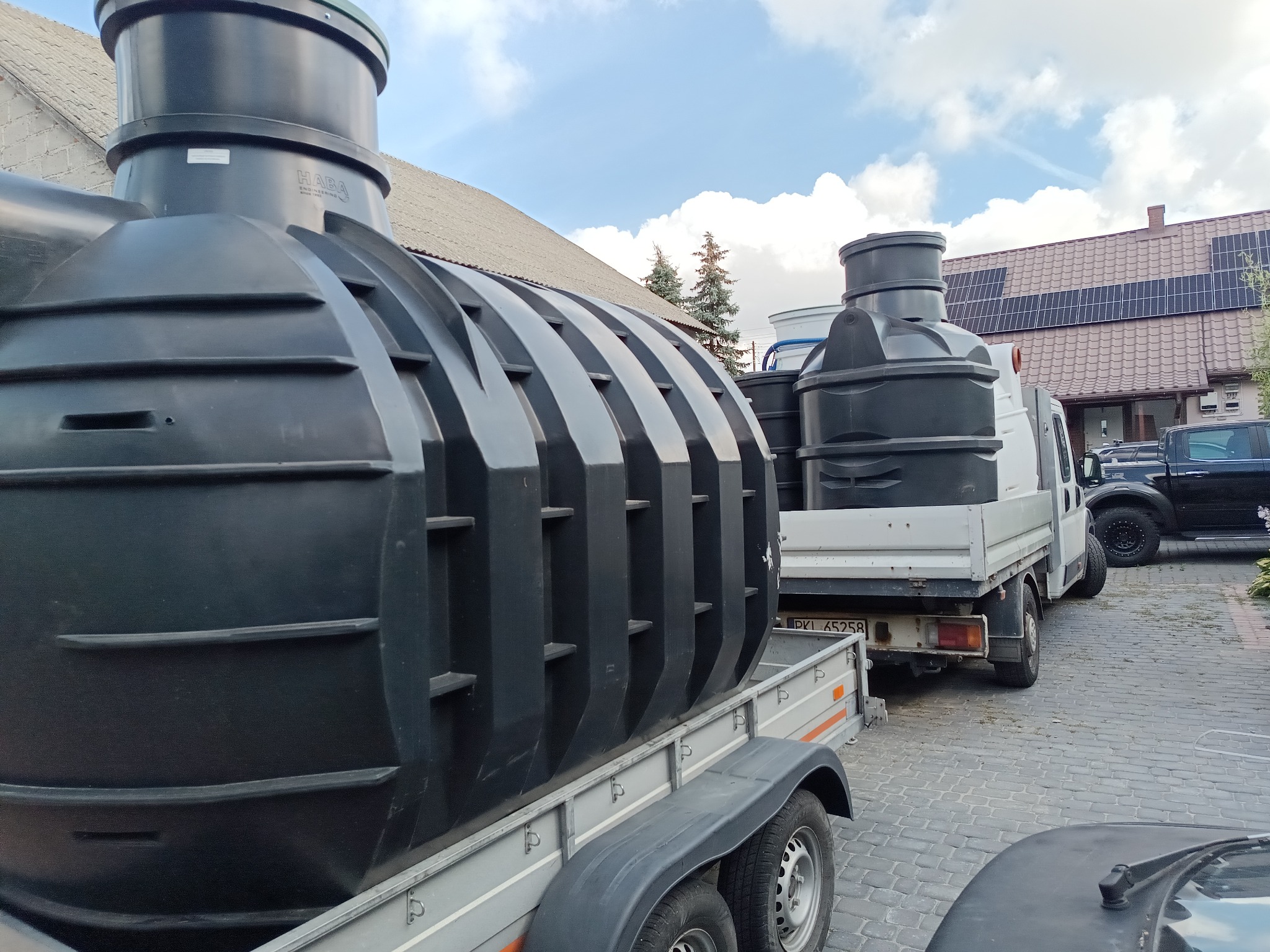Czarne zbiorniki na szarych przyczepach transportowane na posesję z kostki brukowej, w tle budynek z panelami słonecznymi i błękitne niebo z chmurami.