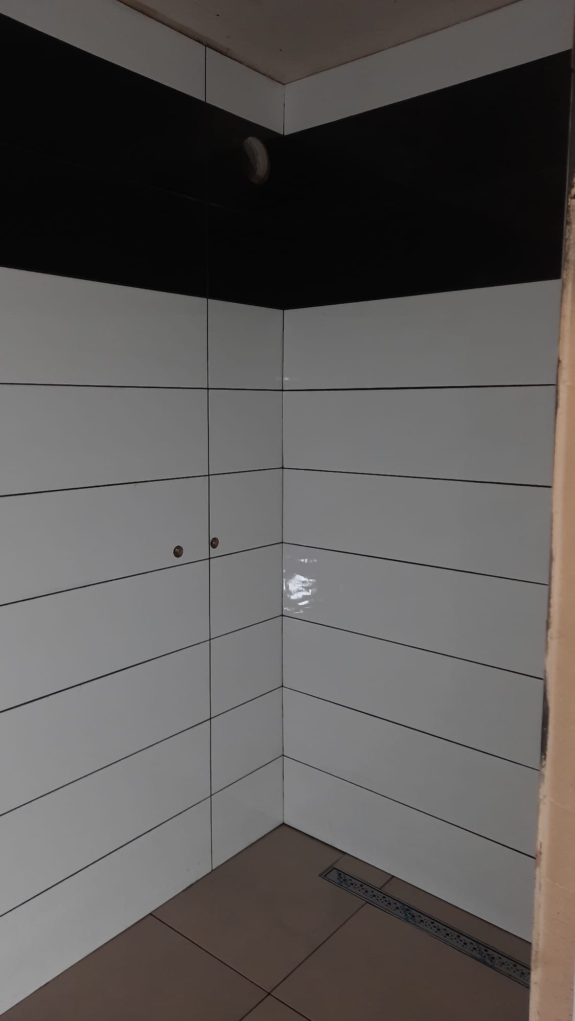 Wykończona łazienka z białymi, poziomymi płytkami i czarnym pasem u góry. Widoczny odpływ liniowy w podłodze i dwa punkty instalacyjne na ścianie.