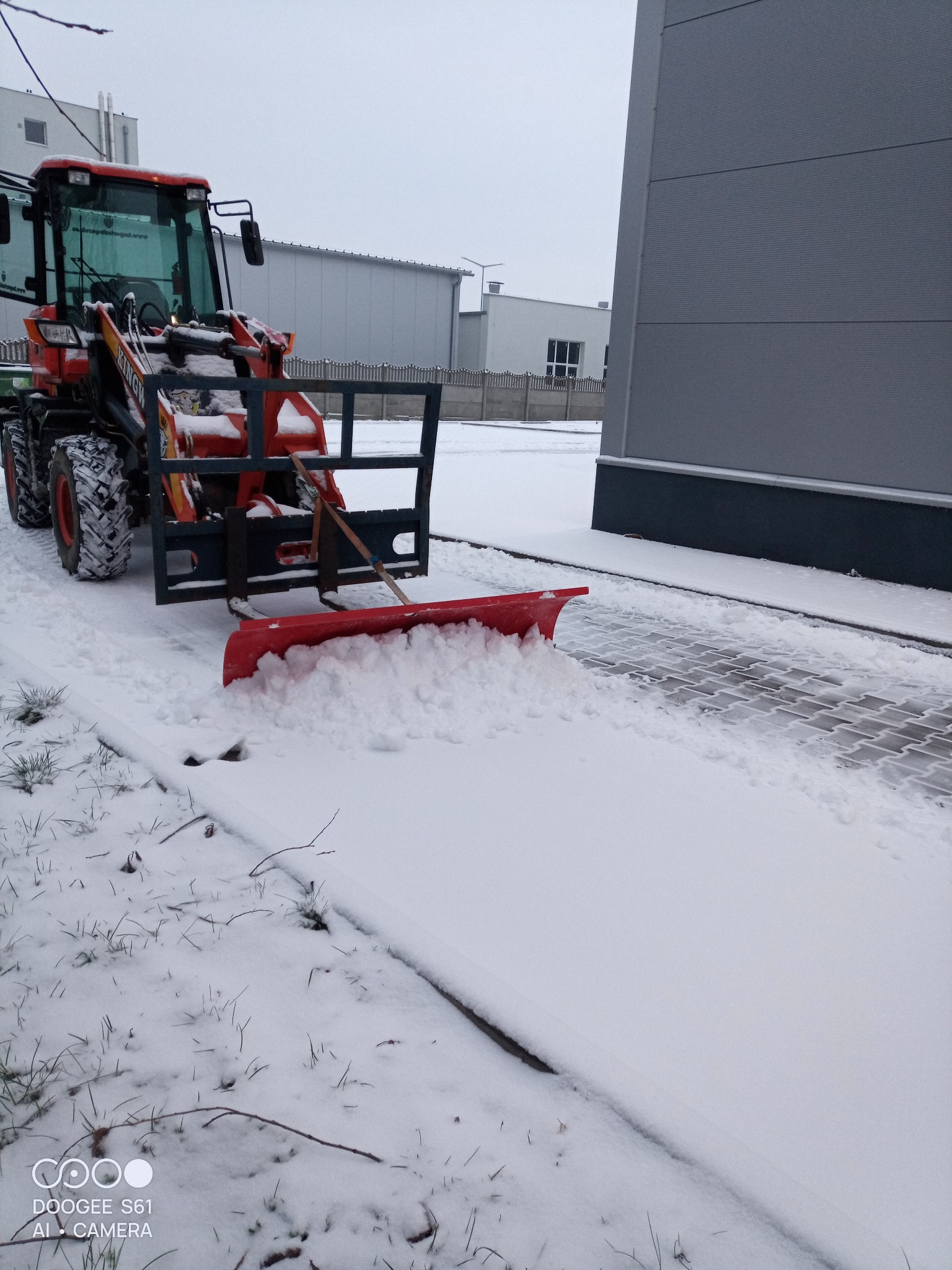 Czerwony traktor z pługiem odśnieża chodnik z kostki brukowej przy szarym budynku w pochmurny dzień. Widoczne ślady opon i odśnieżony fragment.