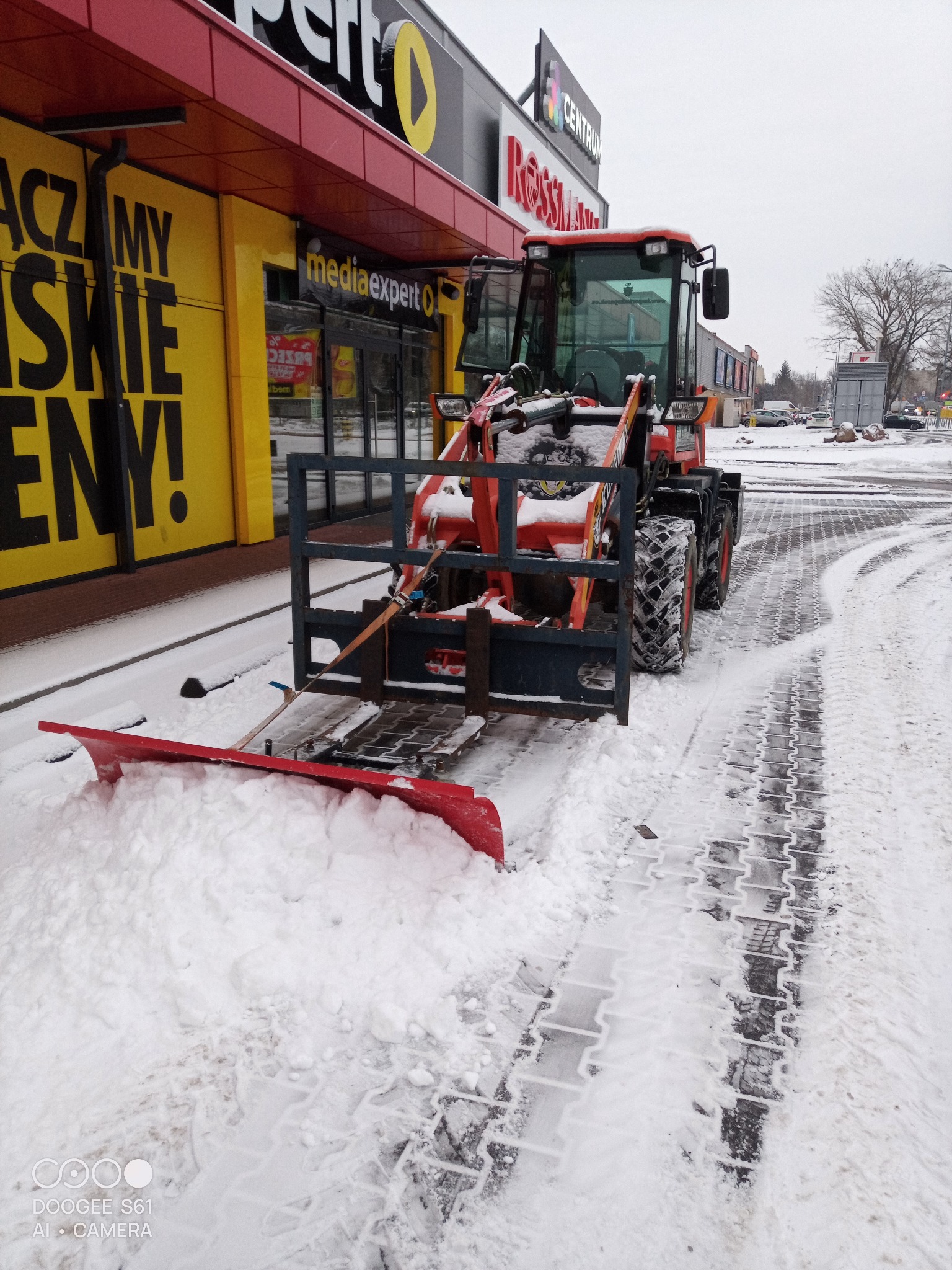 Pomarańczowy traktor z pługiem odśnieża chodnik przed sklepem Media Expert w zimowy dzień. Ślady opon na śniegu.