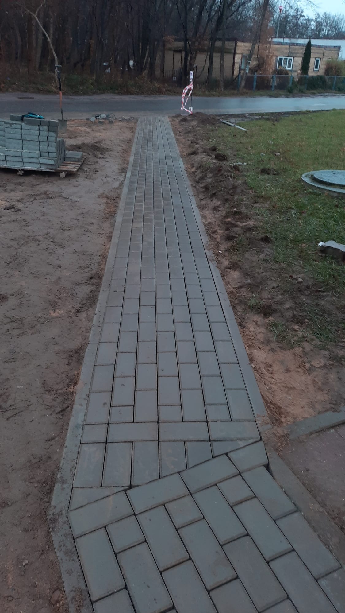 Ułożona ścieżka z szarej kostki brukowej, widok z góry. Część kostki ułożona pod kątem, obok trawnik i droga. Na drugim planie budynek i drzewa.