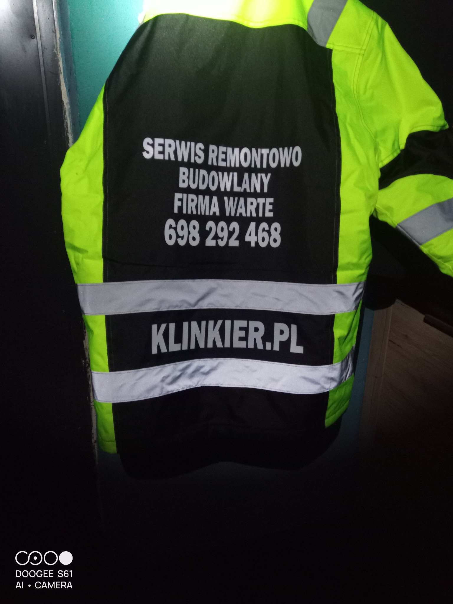 Jaskrawa kamizelka odblaskowa z nadrukiem 'Serwis Remontowo Budowlany Firma Warte' i numerem telefonu, wisząca na drzwiach. Widoczny adres strony internetowej 'klinkier.pl'.