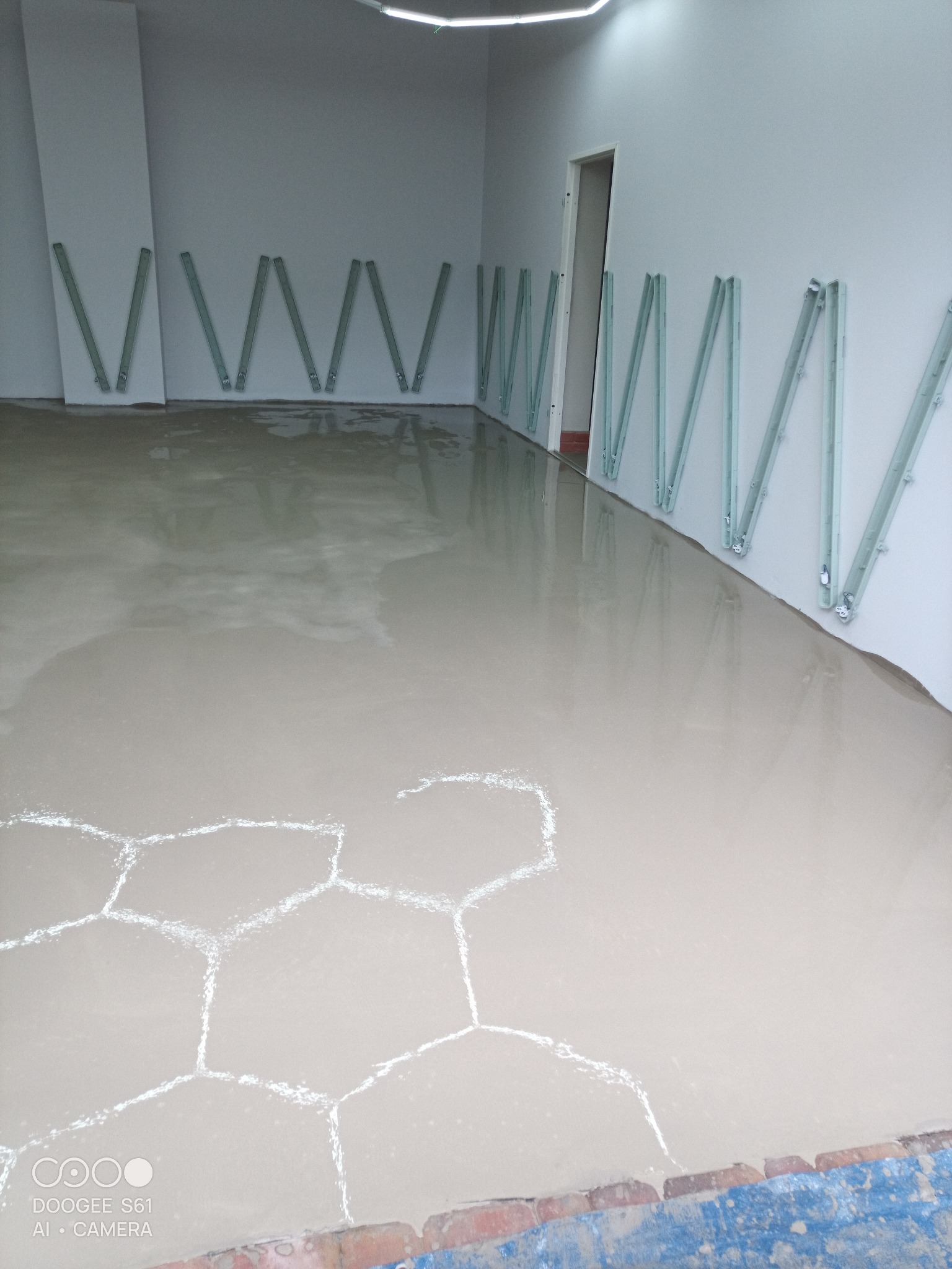 Wylany, jasny podkład podłogowy w pomieszczeniu z białymi ścianami. Na ścianach widoczne pionowe, metalowe listwy. Na podłodze wzór w kształcie plastra miodu.