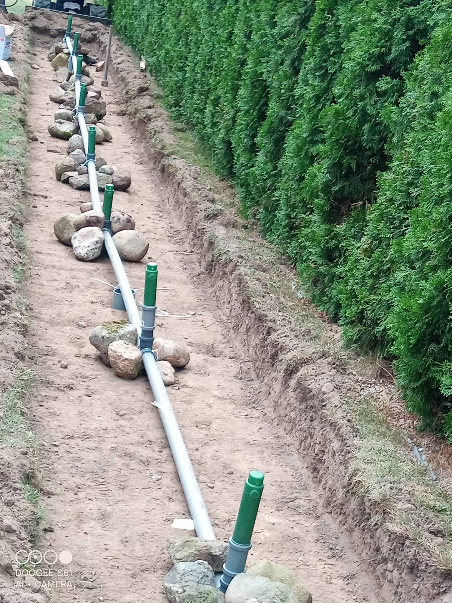 Instalacja drenażowa z rur PVC i kamieni w wykopie, obok żywopłotu. Widok z góry, ukazujący etap prac przy systemie odprowadzania wody.