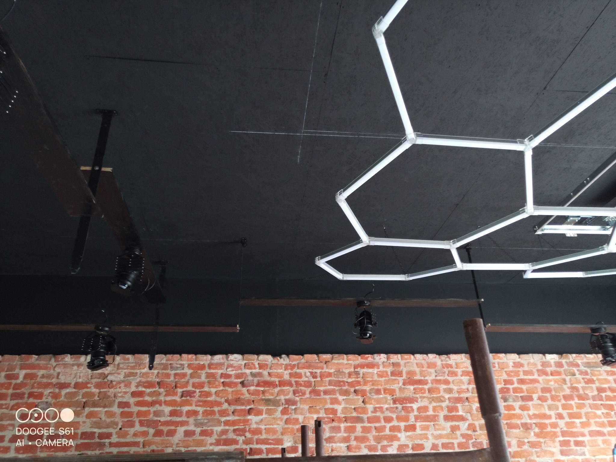 Czarne wnętrze z ceglaną ścianą, oświetlone lampami w kształcie plastra miodu i reflektorami. Widoczne drewniane belki i sufit z widoczną fakturą.