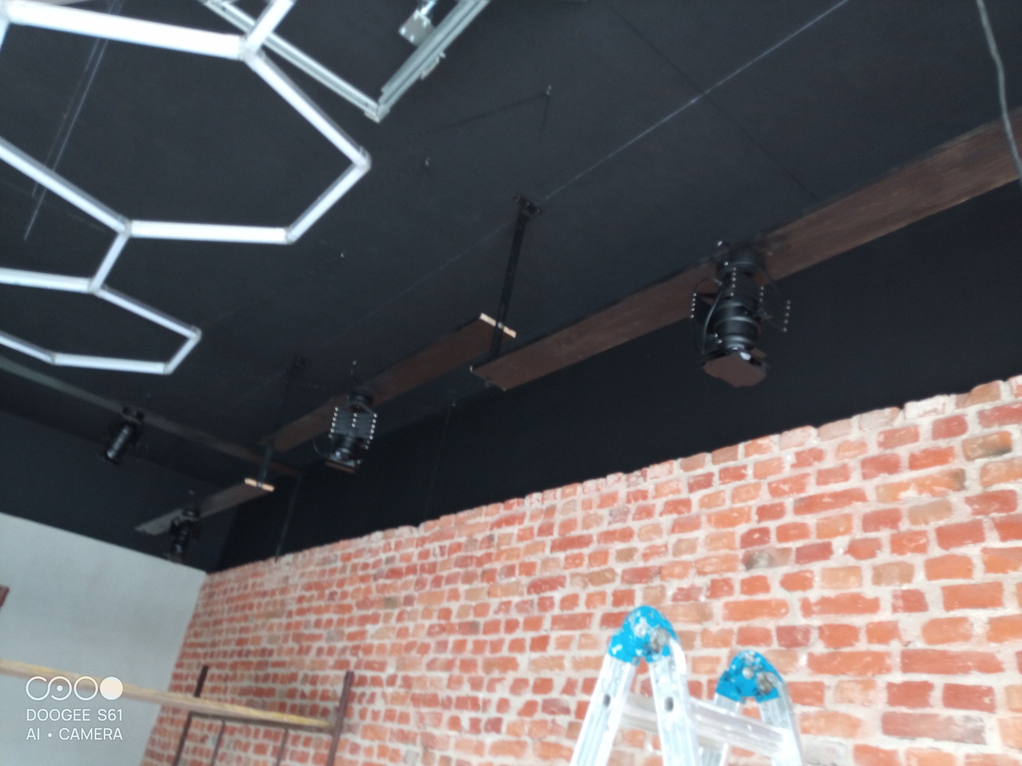 Montaż oświetlenia w biurze w Kole: czarny sufit z lampami na drewnianych belkach, ceglana ściana i drabina. Prace wykończeniowe w toku.