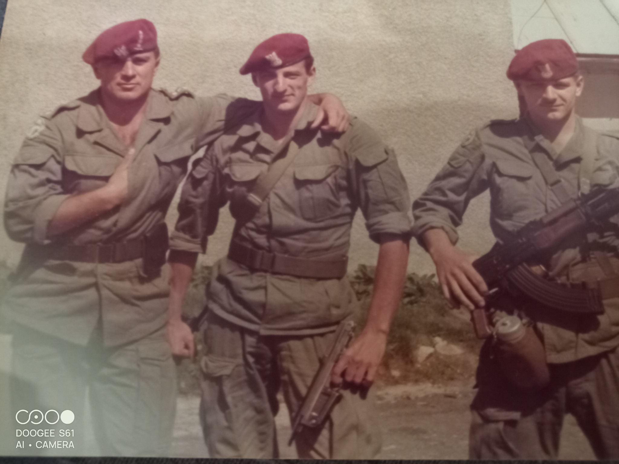 Trzech żołnierzy w mundurach i bordowych beretach, jeden z karabinem AK-47, drugi z pistoletem, pozują na tle jasnej ściany. Zdjęcie ma ciepłą tonację i wygląda na stare.