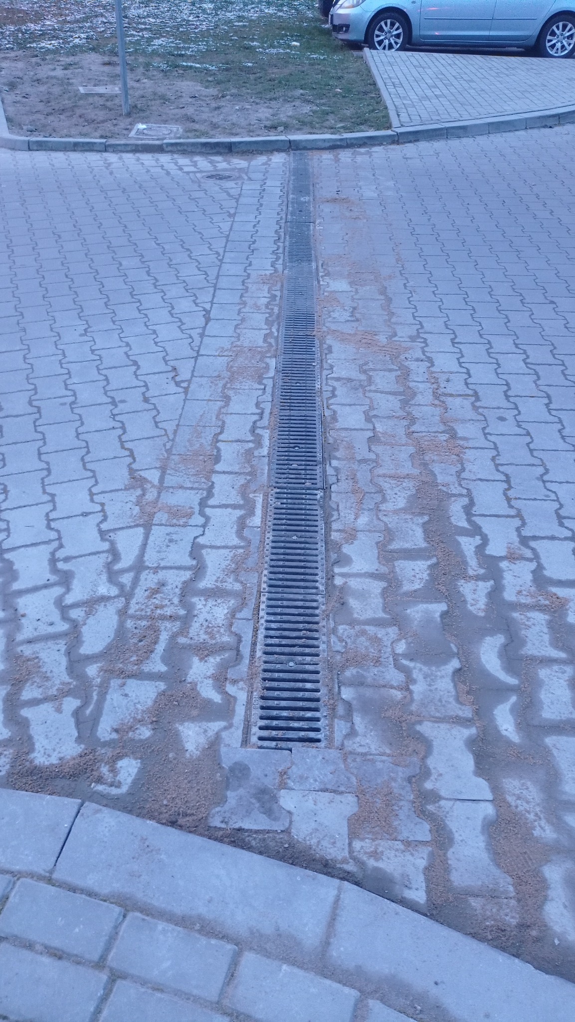 Ułożona, długa, metalowa kratka odpływowa na szarym chodniku z kostki brukowej. Widoczne ślady piasku i wody. W tle fragment trawnika i samochodu.
