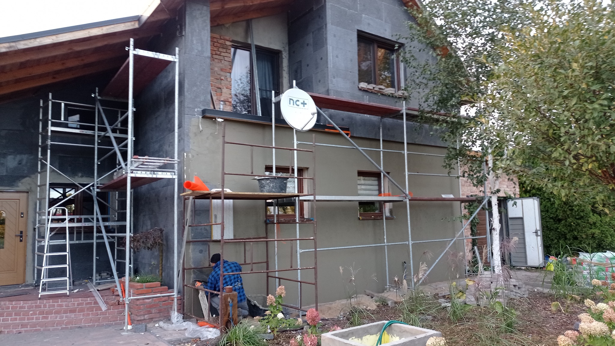 Dom w trakcie ocieplania: szare płyty izolacyjne na elewacji, rusztowania, pracownik w niebieskiej koszuli. Widoczna antena satelitarna i fragment ogrodu.