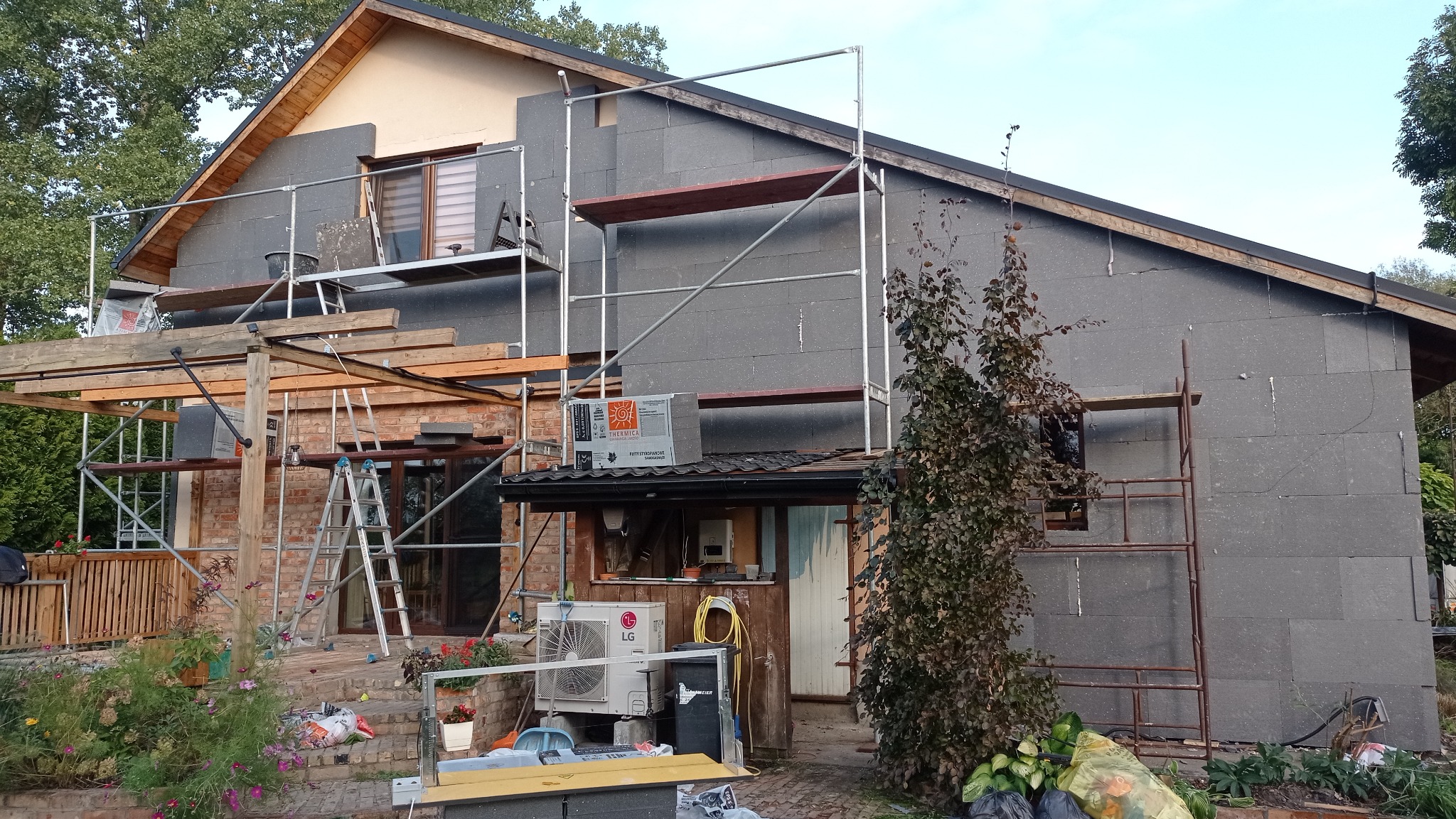 Dom w trakcie ocieplania elewacji styropianem grafitowym, widoczne rusztowania, materiały budowlane i klimatyzator LG na zewnątrz budynku.