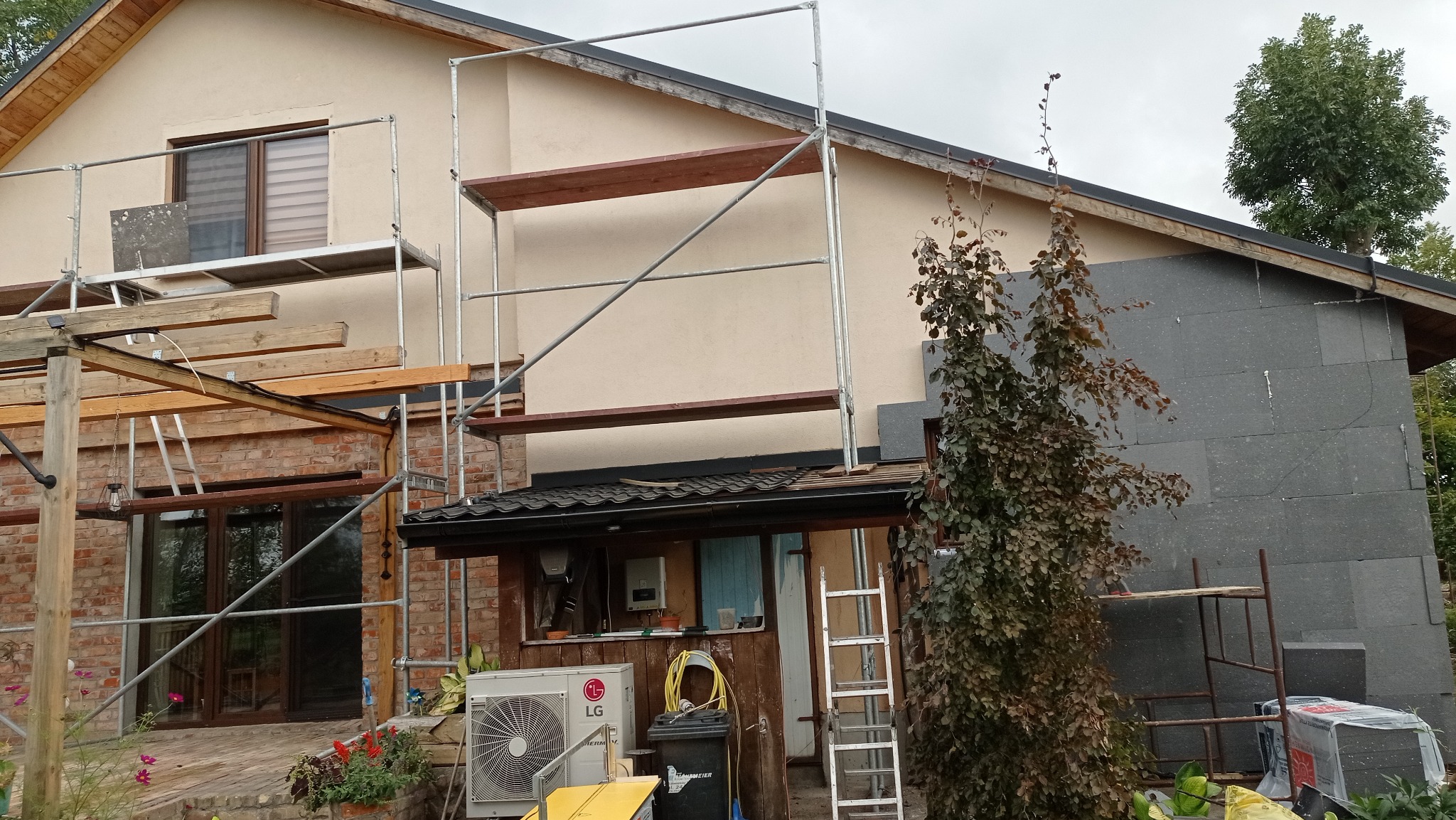 Dom w trakcie ocieplania elewacji: fragment ściany z szarym styropianem, rusztowania, widoczny klimatyzator LG i drzewo z bordowymi liśćmi.