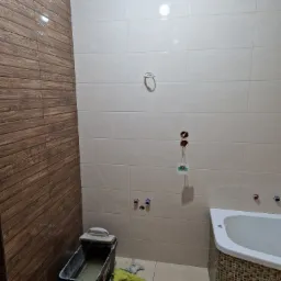 Łazienka w trakcie prac wykończeniowych: ściana z płytek imitujących drewno, jasne płytki na pozostałych ścianach, przygotowana wanna z mozaiką, wiadro z zaprawą i narzędzia glazurnicze.