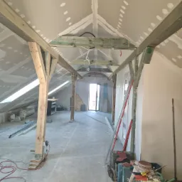 Poddasze w trakcie prac wykończeniowych: widoczne drewniane belki konstrukcyjne, płyty kartonowo-gipsowe na skosach, narzędzia budowlane i drabina na podłodze, zarys okna dachowego w tle.