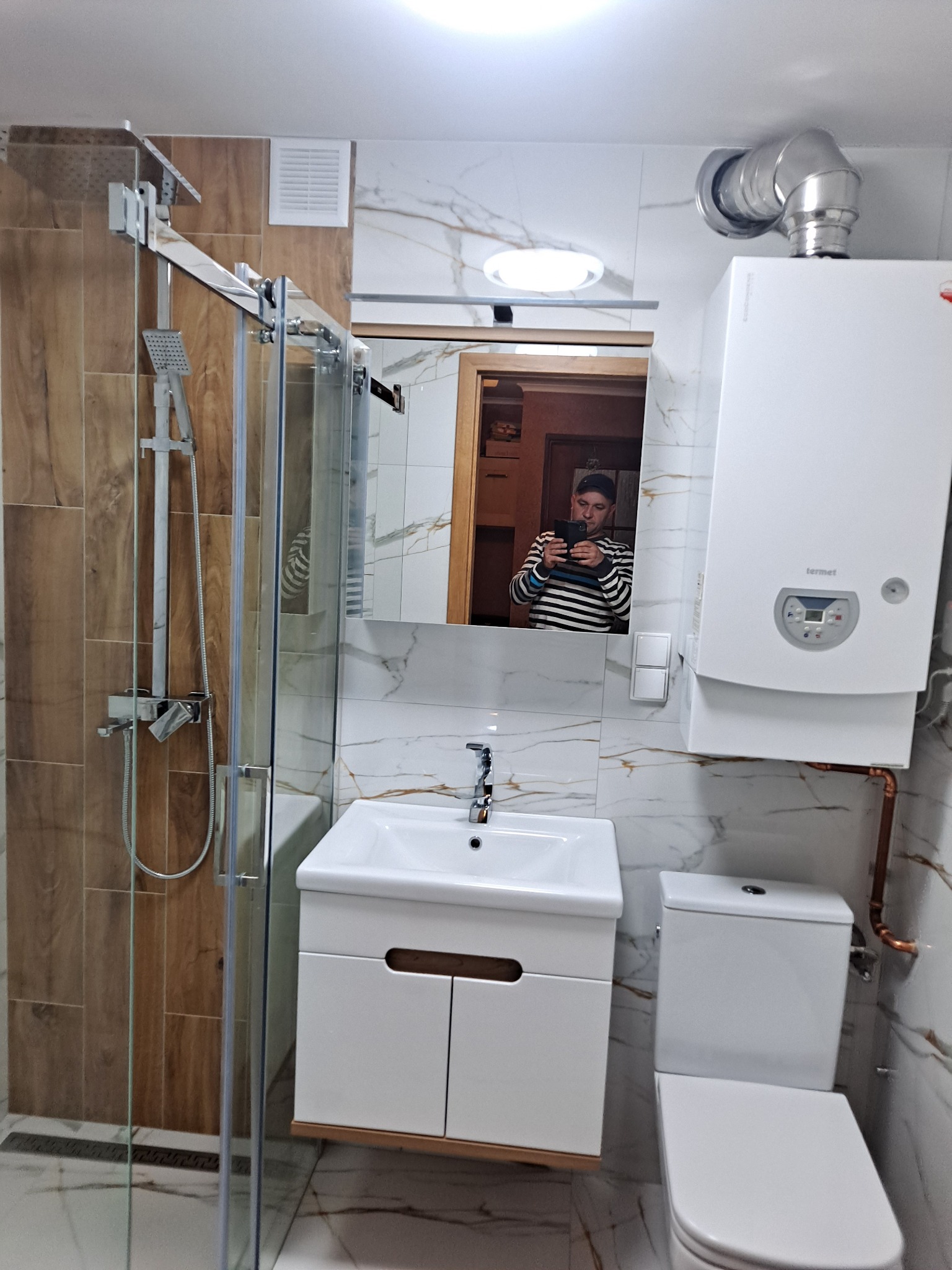 Nowoczesna łazienka po remoncie: kabina prysznicowa z deszczownicą, umywalka z szafką, toaleta i piec gazowy Termet na tle marmurowych płytek, widoczne odbicie fotografa w lustrze.