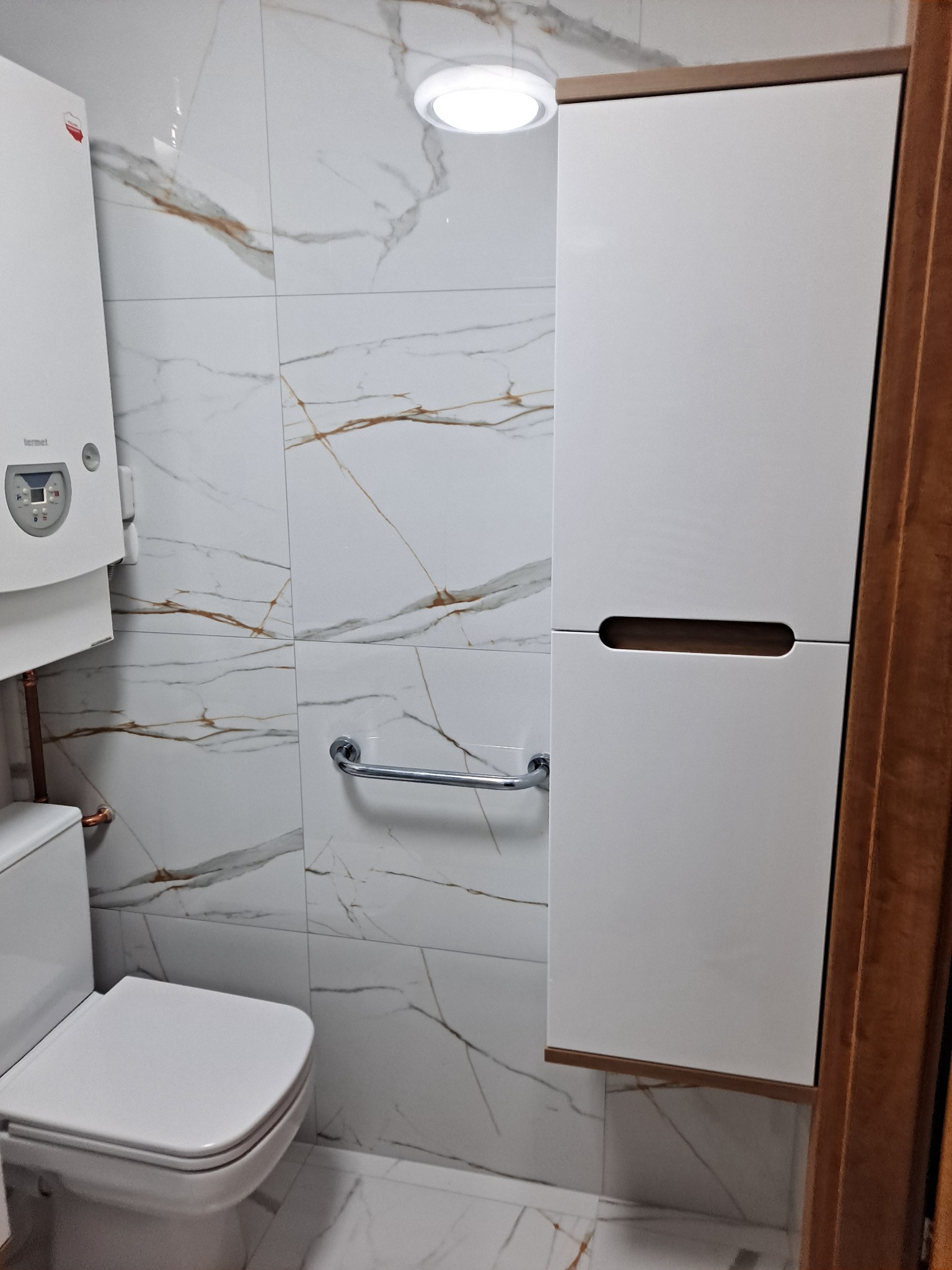 Biała toaleta z wiszącą szafką i grzejnikiem na ścianie z marmurowymi płytkami w łazience po remoncie.