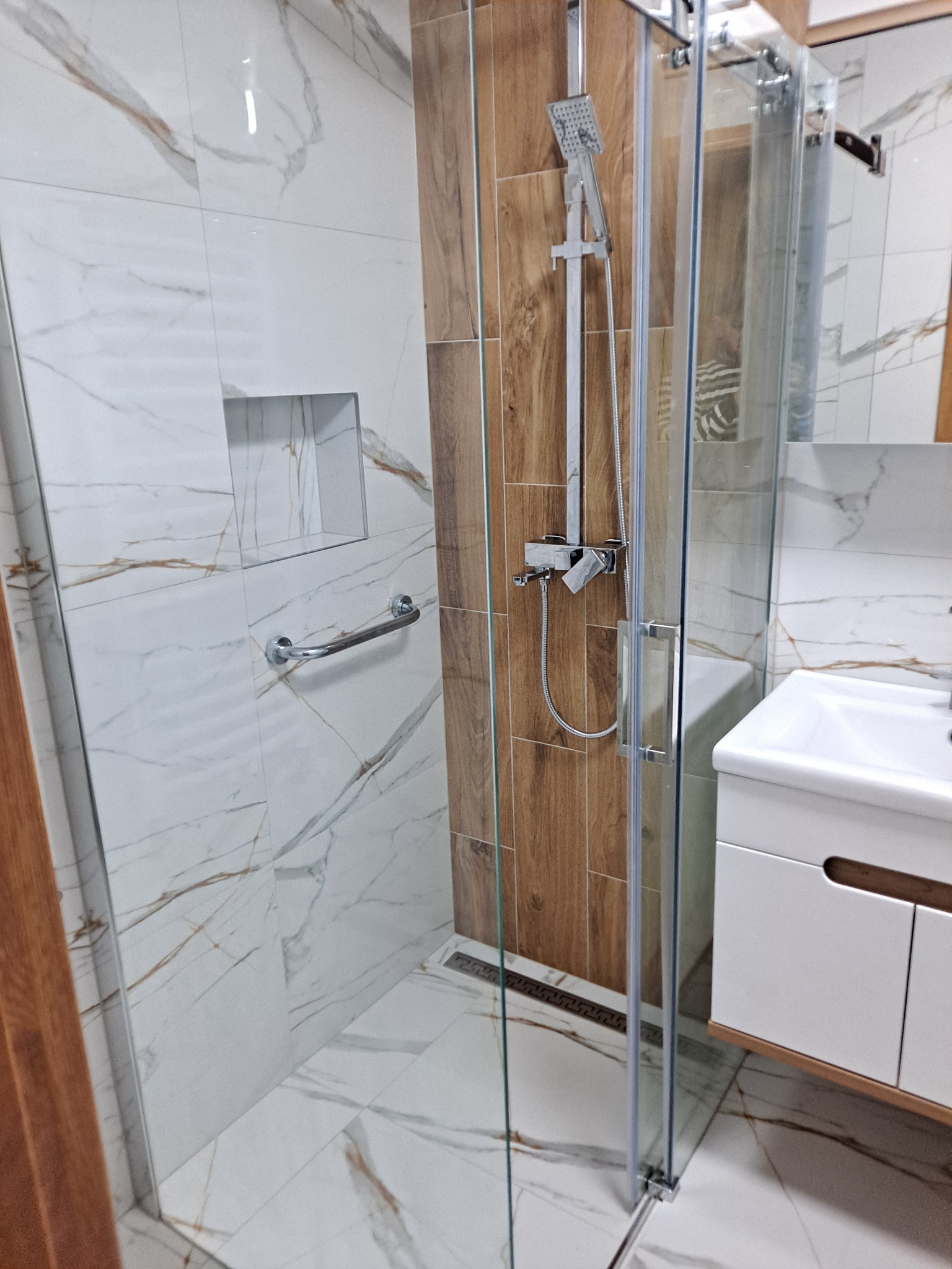 Nowoczesna kabina prysznicowa z chromowaną armaturą, wykończona płytkami imitującymi marmur i drewno, z widoczną wnęką w ścianie i uchwytem dla niepełnosprawnych.