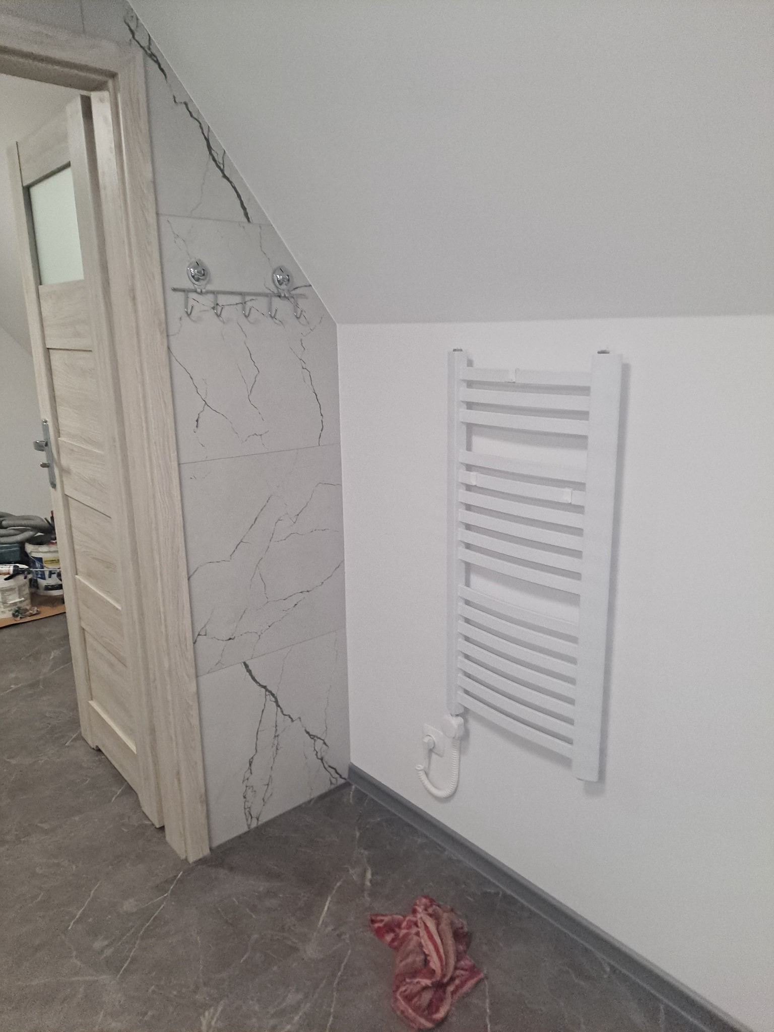 Wykończona łazienka na poddaszu: biały grzejnik drabinkowy na białej ścianie, płytki imitujące marmur, drewniane drzwi, szara podłoga imitująca kamień, oraz wieszak z haczykami.