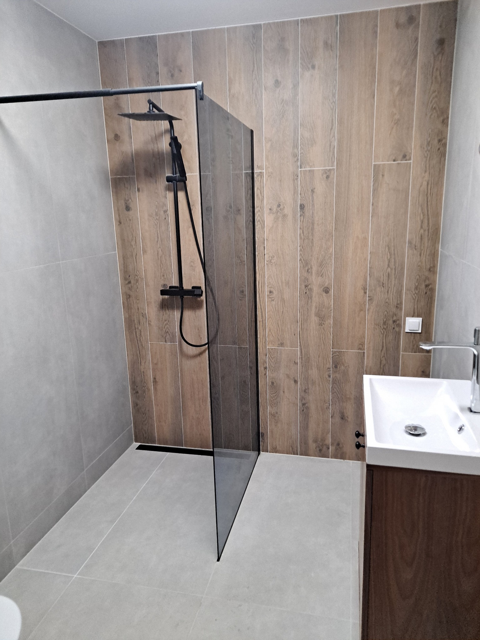 Nowoczesna łazienka z kabiną typu walk-in, drewnopodobnymi płytkami na ścianie i szarą armaturą, minimalistyczna umywalka z szafką.