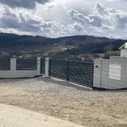 Nowoczesna brama wjazdowa z poziomych, grafitowych profili, osadzona na betonowych słupkach i murze z szarej kostki, na tle pagórkowatego krajobrazu pod pochmurnym niebem.