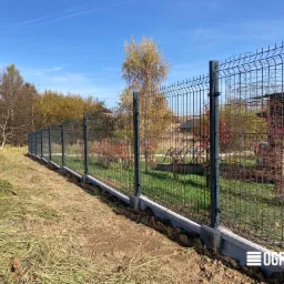 Długie, szare ogrodzenie panelowe z podmurówką betonową, zamontowane na trawiastym terenie z widocznym budynkiem w tle, sfotografowane w słoneczny dzień.