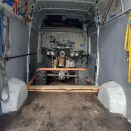 Quad zabezpieczony pasami transportowymi na drewnianej palecie wewnątrz białego busa dostawczego.