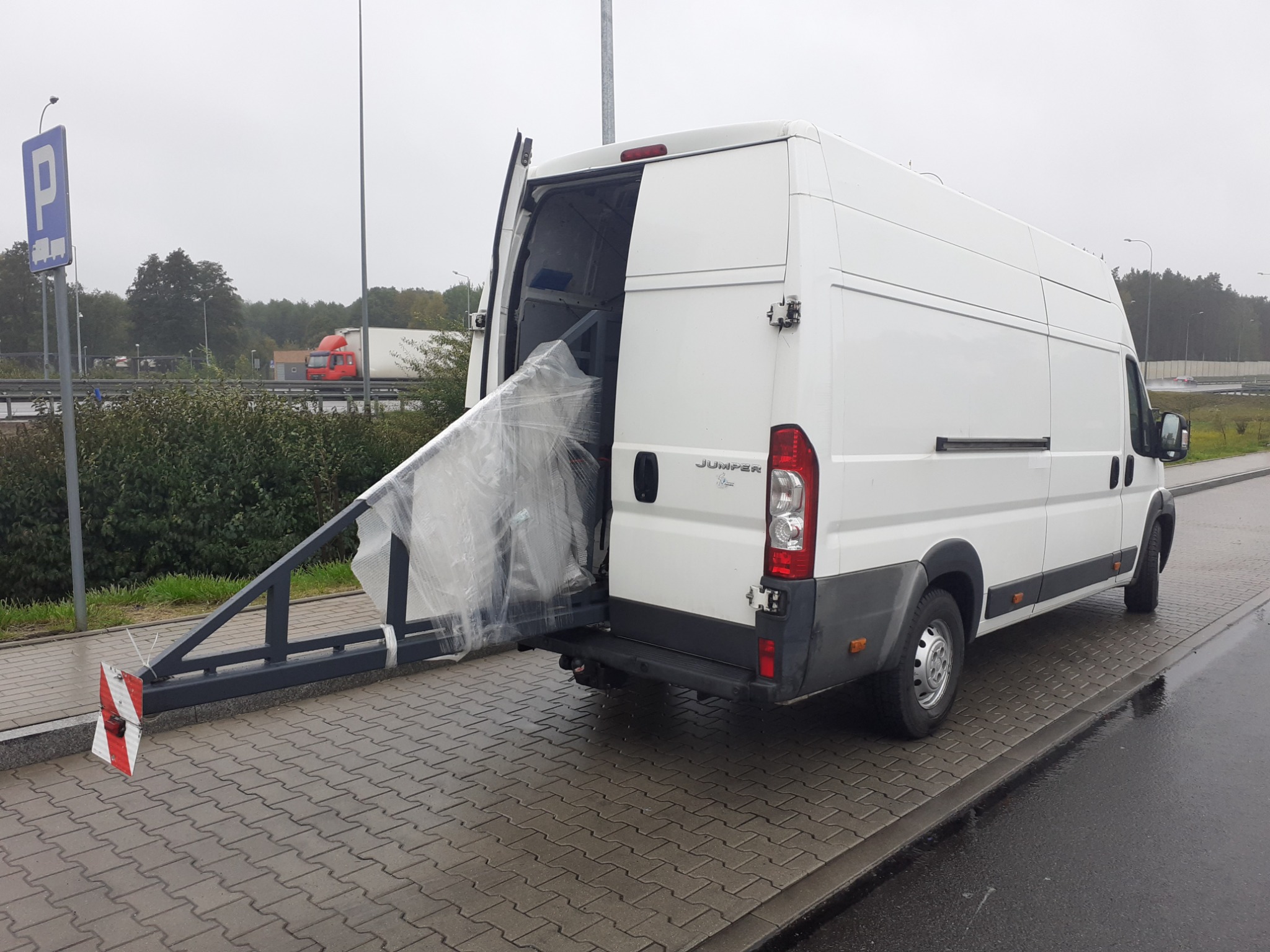 Biały bus z otwartymi drzwiami załadowany długim, zabezpieczonym folią elementem konstrukcyjnym z wystającym stalowym wspornikiem z oznaczeniami ostrzegawczymi na końcu, zaparkowany na kostce...