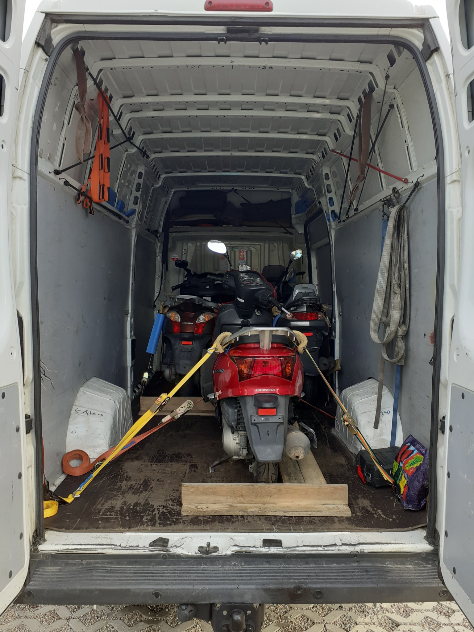 Wnętrze białego busa z trzema skuterami Honda zabezpieczonymi pasami transportowymi i drewnianymi klockami, widok z tyłu pojazdu.