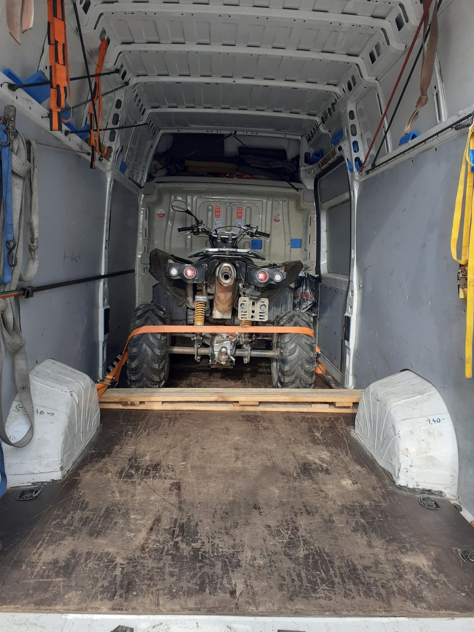 Quad zabezpieczony pasami transportowymi na drewnianej palecie wewnątrz białego busa dostawczego.