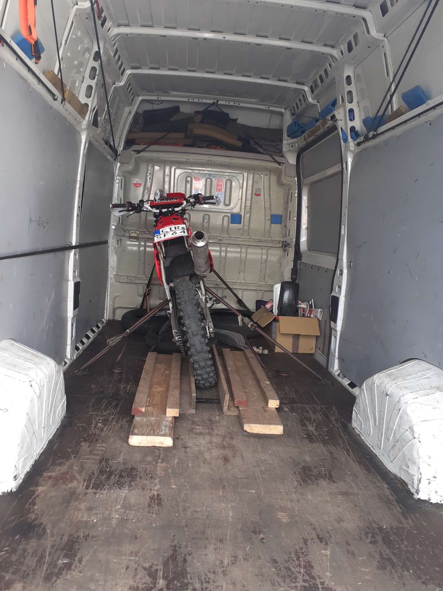 Wnętrze białego busa z zabezpieczonym motocyklem crossowym na drewnianych legarach, widoczne pasy mocujące i kartonowe pudła.