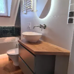 Nowoczesna łazienka na poddaszu z drewnianym blatem, umywalką nablatową, podświetlanym lustrem i grzejnikiem drabinkowym.