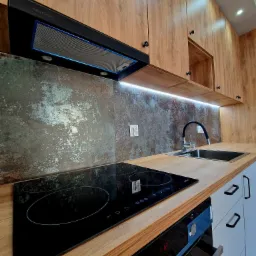 Nowoczesna kuchnia z drewnianymi szafkami, blatem i czarną płytą indukcyjną, zlokalizowana w Oławie, z metalicznym panelem nad blatem i podświetleniem LED.