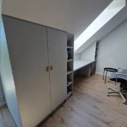 Pokój na poddaszu ze skośnym oknem dachowym, wyposażony w szarą szafę z drewnianymi uchwytami, biurko w zabudowie z półkami i szufladami oraz krzesło biurowe na kółkach.