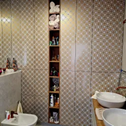 Wnękowa szafa w łazience z półkami na kosmetyki i ręczniki, biało-złote płytki z geometrycznym wzorem, umywalki nablatowe.