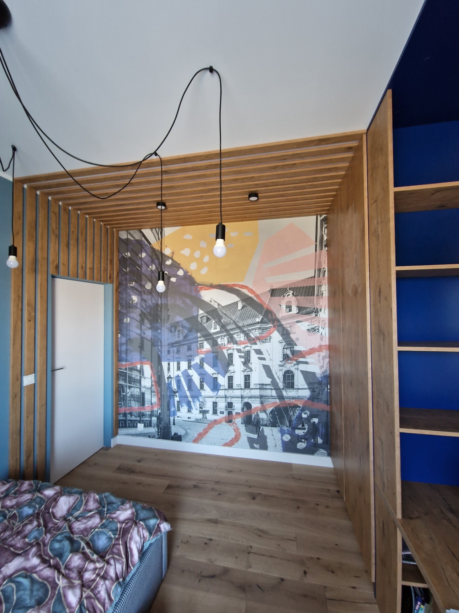 Pokój z drewnianą zabudową, regałami w kolorze niebieskim i fototapetą z motywem miejskim, oświetlony wiszącymi lampami z odsłoniętymi żarówkami.