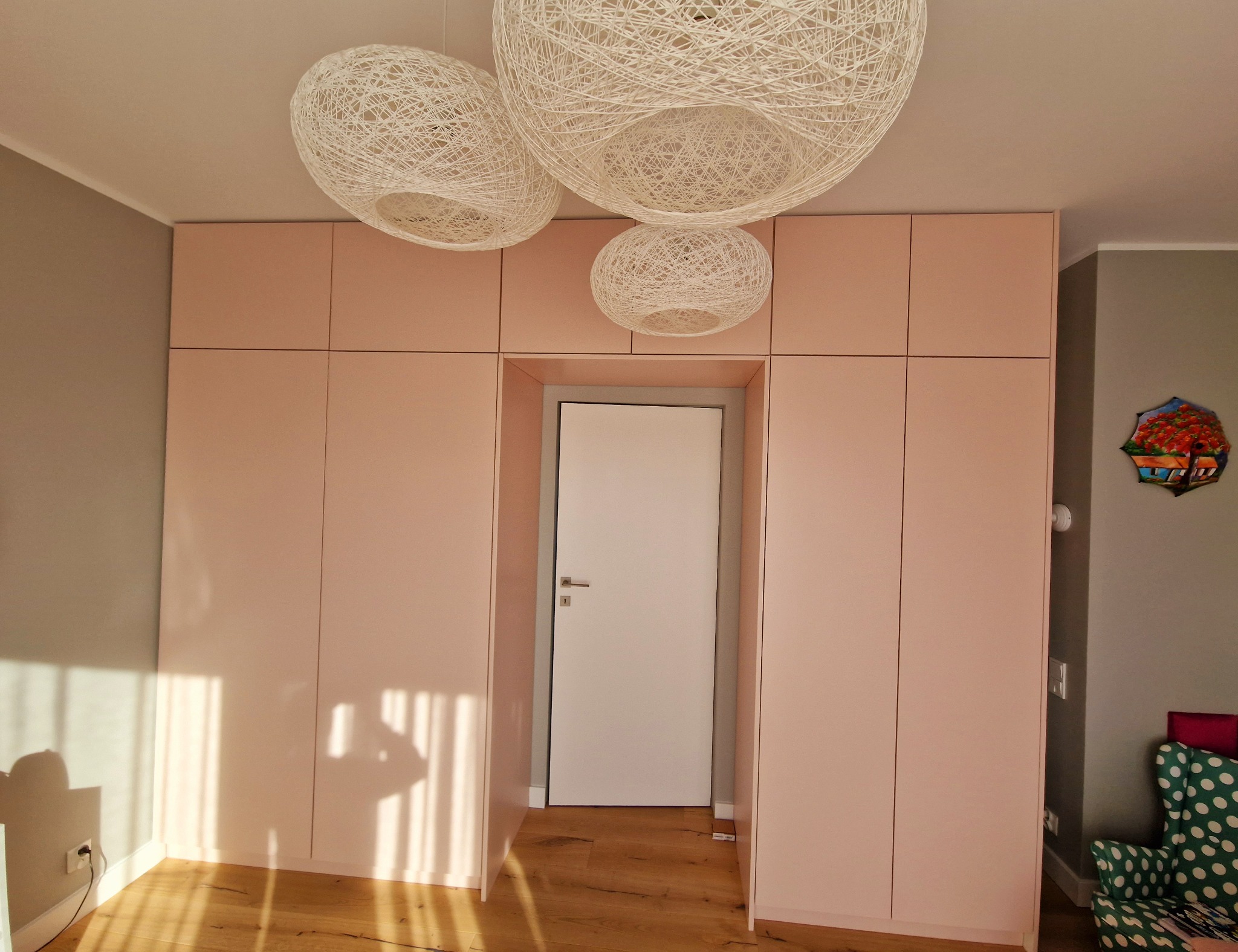 Różowa zabudowa meblowa z szafami i przejściem na drzwi, oświetlona trzema okrągłymi lampami ze sznurka, na podłodze parkiet, wpadające promienie słońca