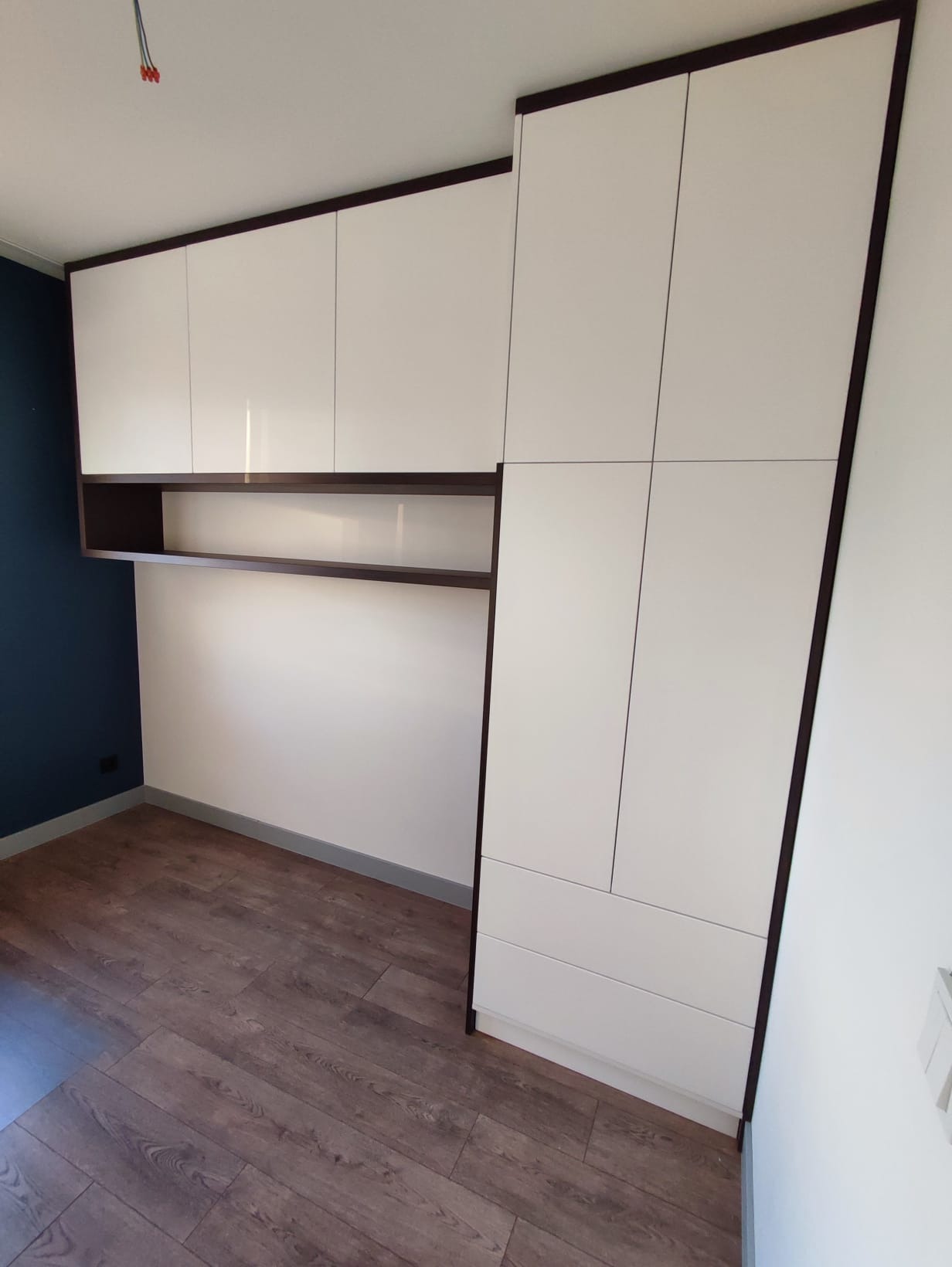 Biała szafa z ciemnobrązowymi obramowaniami, obejmująca szafki wiszące i szuflady, na tle częściowo pomalowanej ściany i paneli podłogowych.