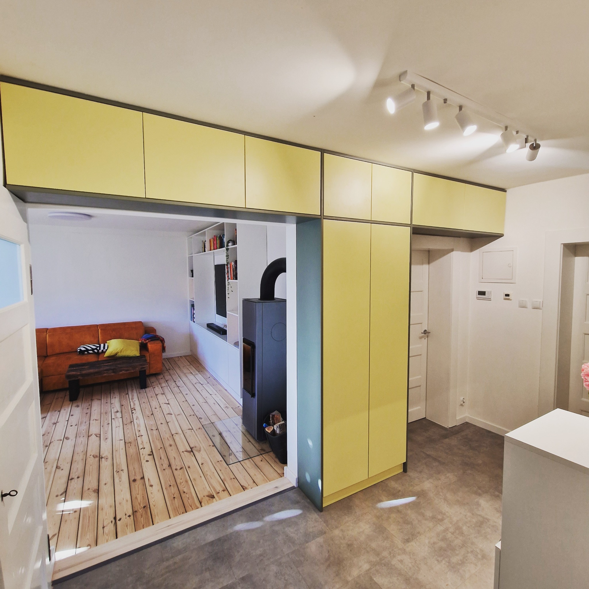 Żółte szafki podwieszane z szarym obramowaniem nad przejściem do salonu z kominkiem i drewnianą podłogą, widok z korytarza z szarą podłogą.