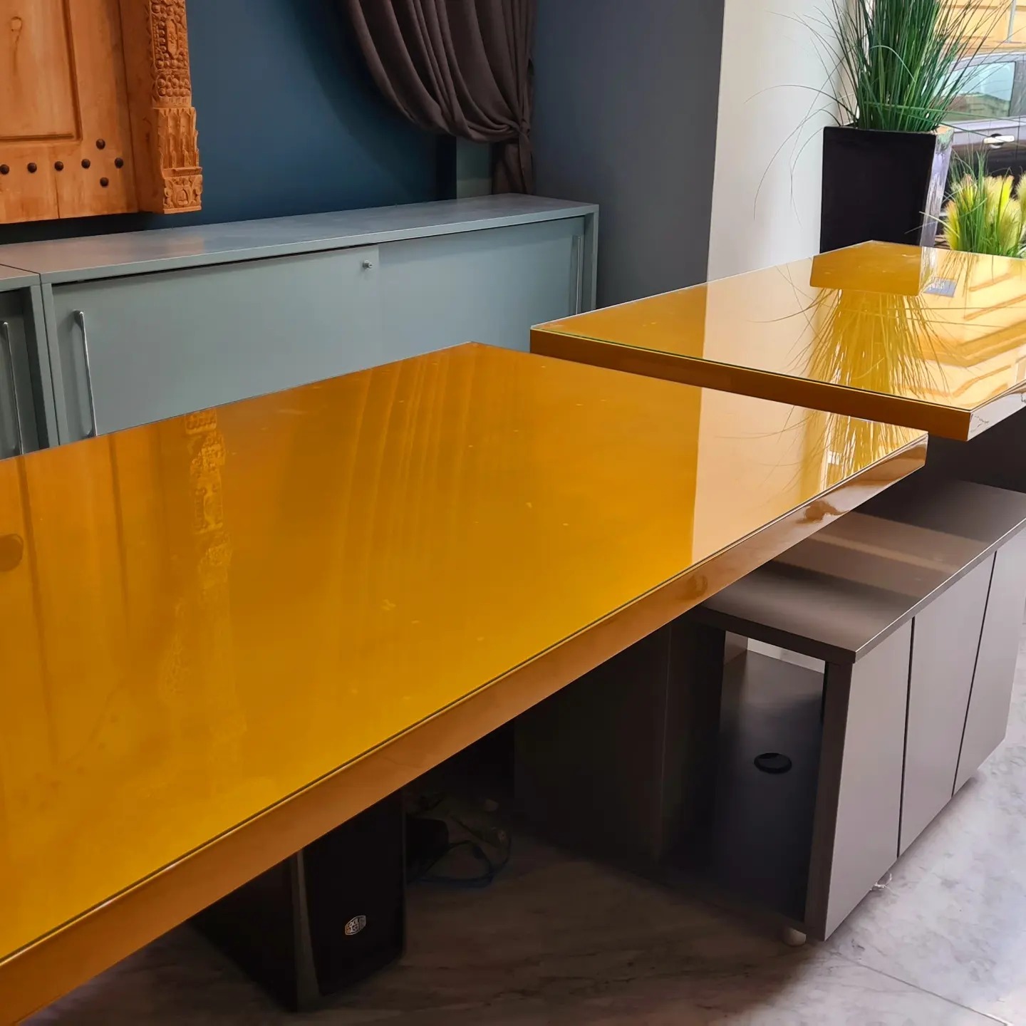 Dwa złote, lakierowane blaty na szafkach z szarymi frontami, w tle dekoracyjna szafka z rzeźbionymi elementami i ciemną zasłoną.