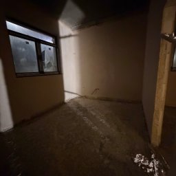 IDB HOUSE - Pokój po nałożeniu tynków: gładkie ściany, widoczne okno z folią ochronną, surowa podłoga. Przygotowanie do dalszych prac wykończeniowych w nowym budownictwie.