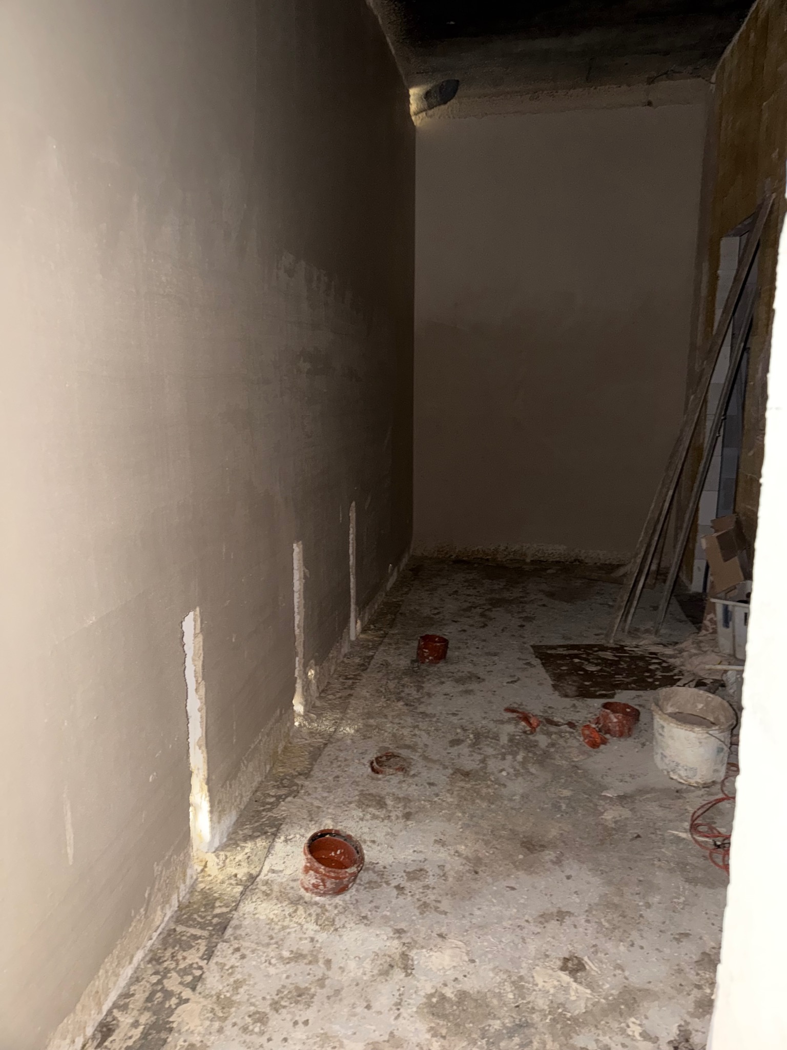 Świeżo położone, jasne tynki na ścianach w wąskim pomieszczeniu. Widoczne pionowe bruzdy w tynku, narzędzia i wiadro na podłodze. Surowy stan wykończenia wnętrza.