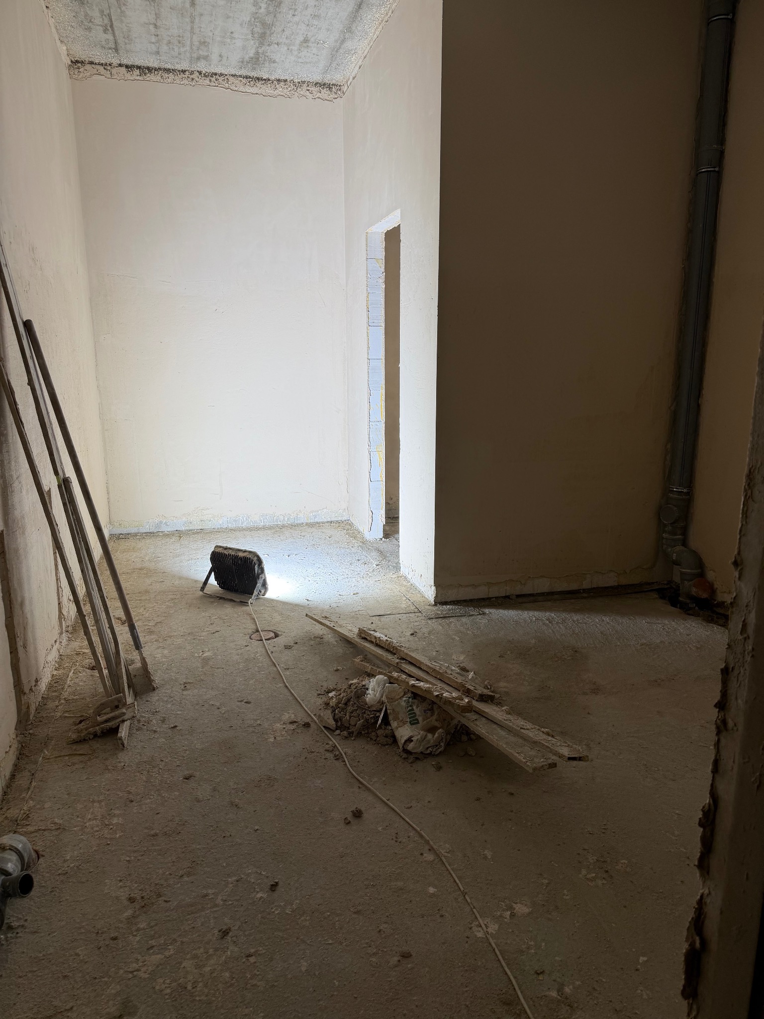 Wnętrze po tynkowaniu maszynowym: gładkie ściany, widoczny sprzęt budowlany i resztki materiałów na podłodze. Widoczne rury i oświetlenie robocze.