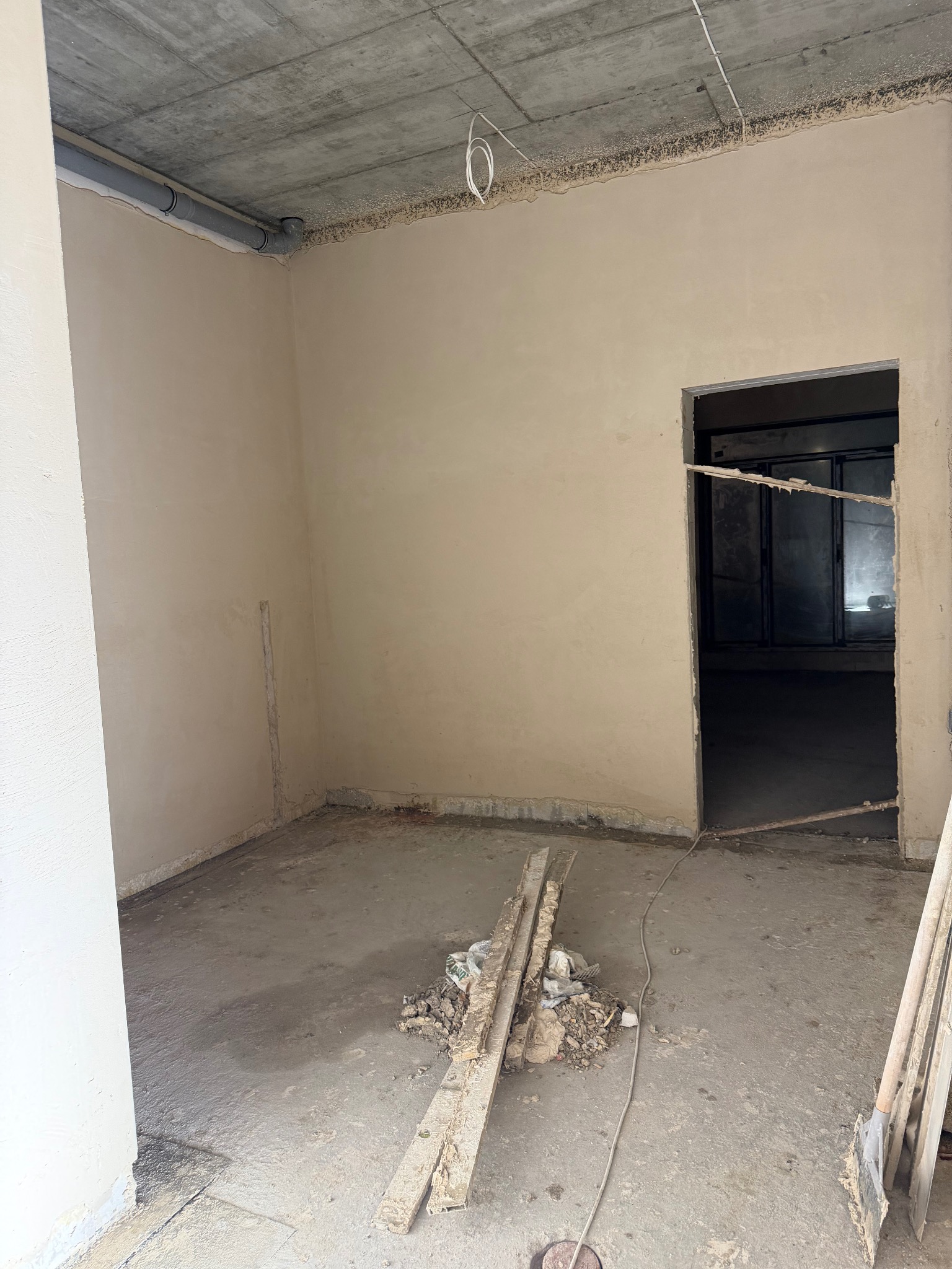 Wnętrze po tynkowaniu maszynowym: gładkie, jasne ściany, surowy betonowy sufit z instalacją, widoczne narzędzia i resztki materiałów na podłodze. Przygotowanie do dalszych prac wykończeniowych.