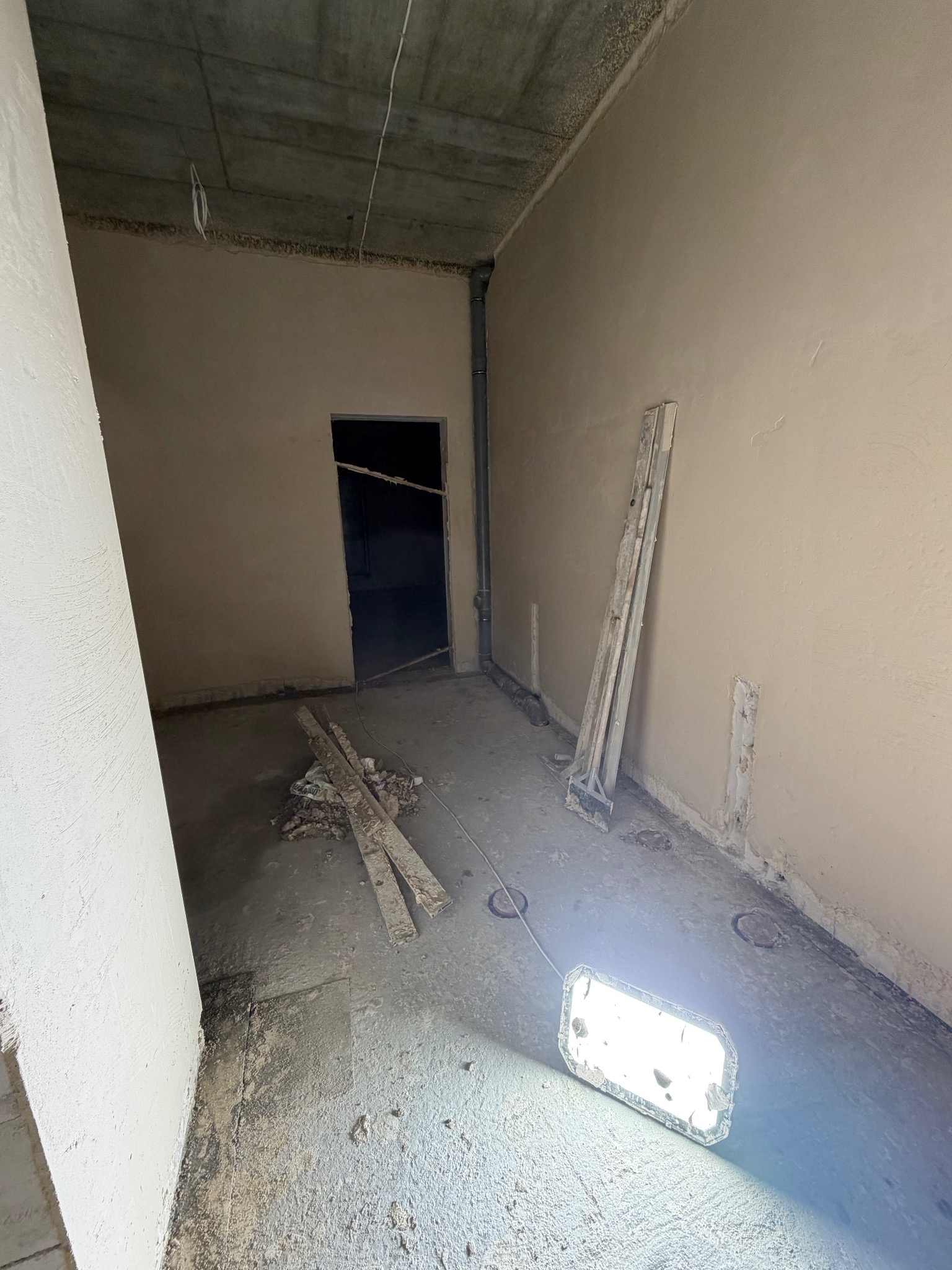 Wnętrze w trakcie prac wykończeniowych: świeżo otynkowane ściany, surowy betonowy strop, narzędzia i lampa budowlana na podłodze. Widoczne ślady po instalacjach.