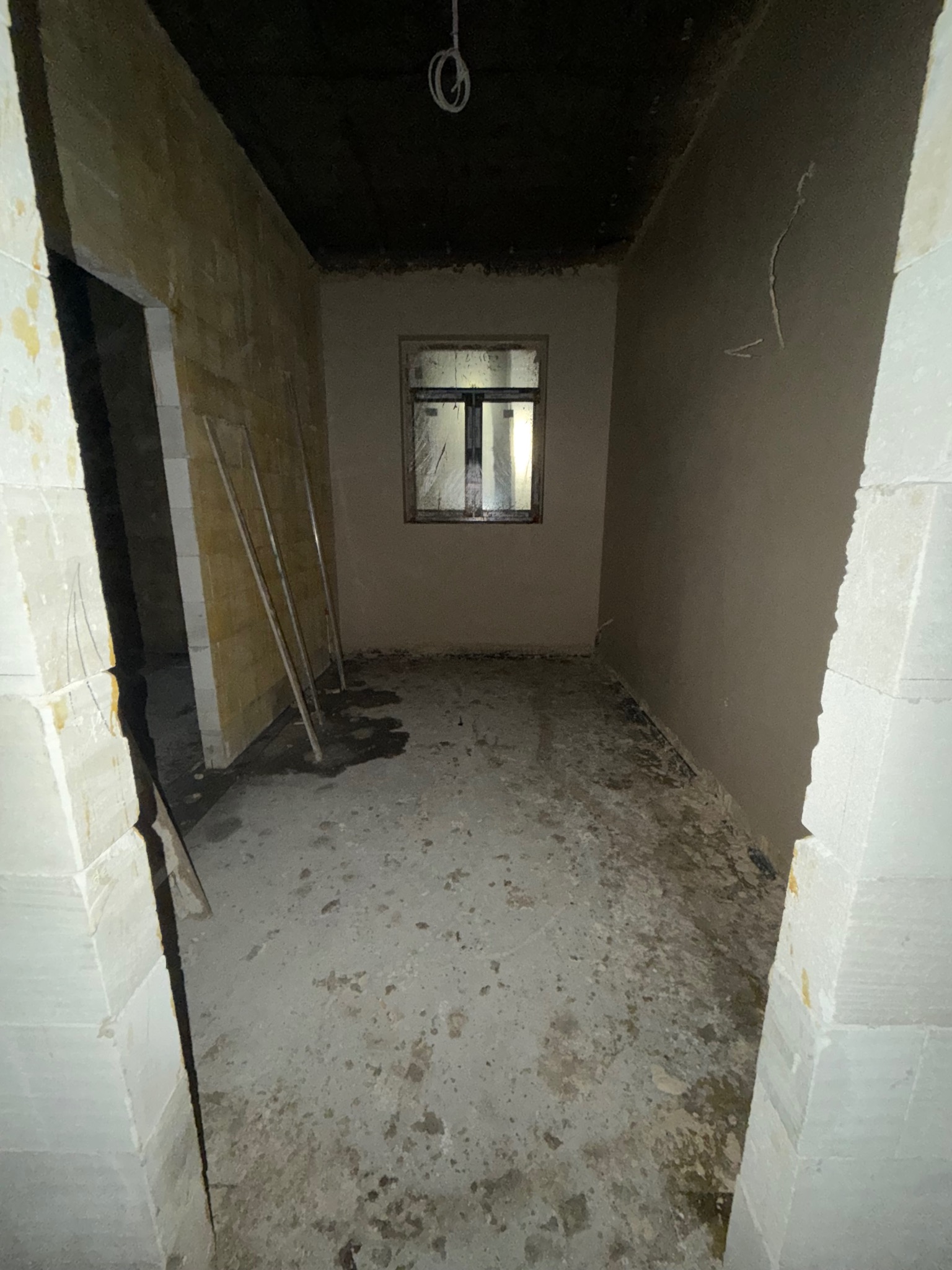 Pokój w trakcie prac wykończeniowych: częściowo otynkowane ściany, widoczne profile, surowy strop, okno z widokiem na zewnątrz. Wnętrze w stanie surowym.