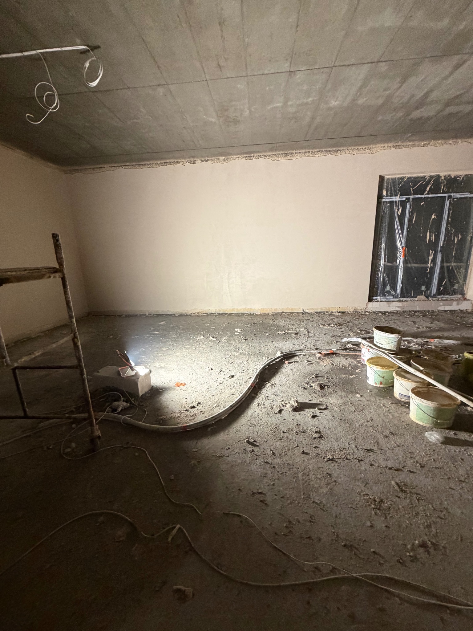 Wnętrze w trakcie prac tynkarskich: ściany częściowo pokryte tynkiem, widoczne puszki z farbą, kable, drabina i prowizoryczne oświetlenie. Okno zabezpieczone folią.